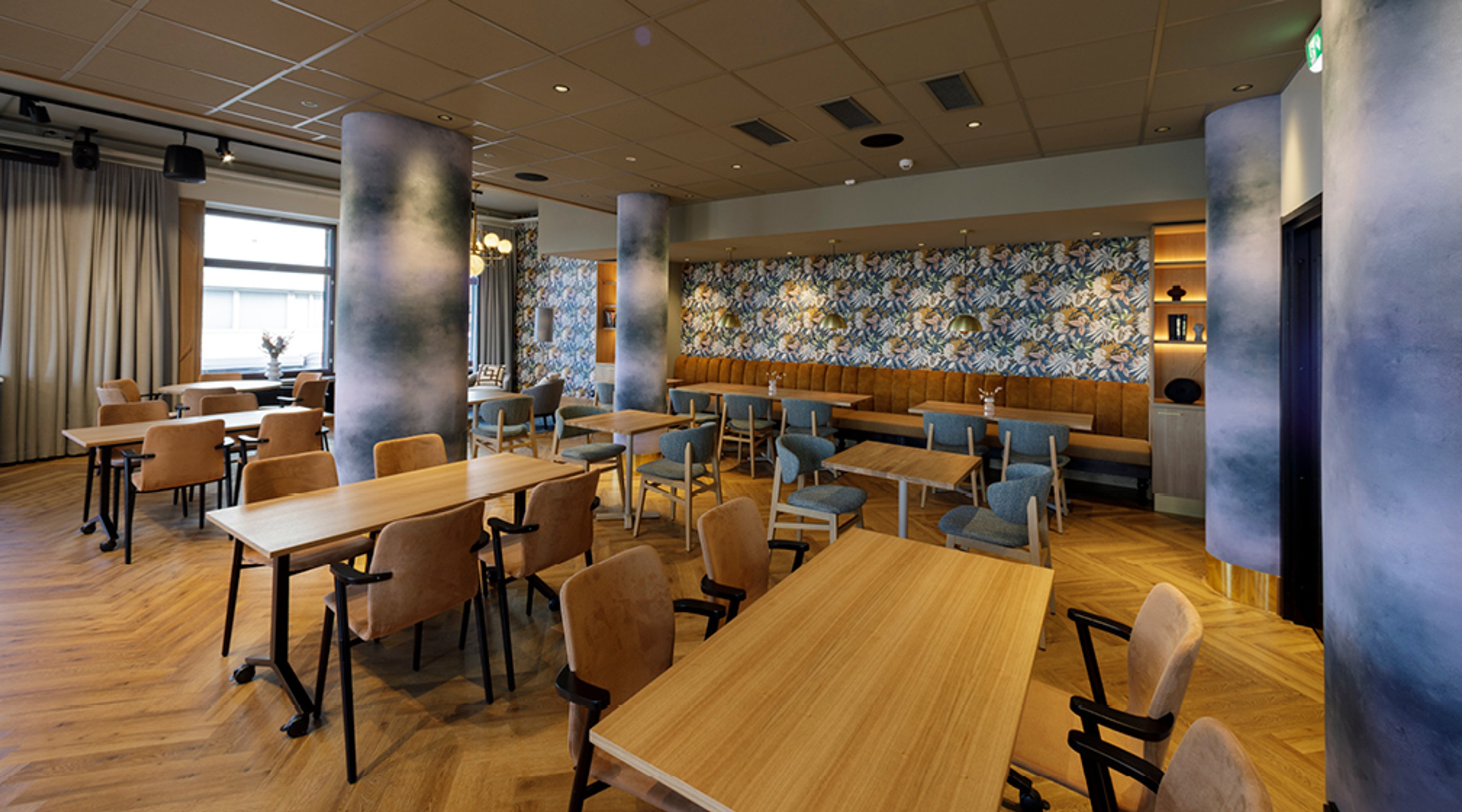 Modern restaurang med träbord, blå och bruna stolar samt blommig tapet på väggen. Rummet har stora fönster och trägolv.