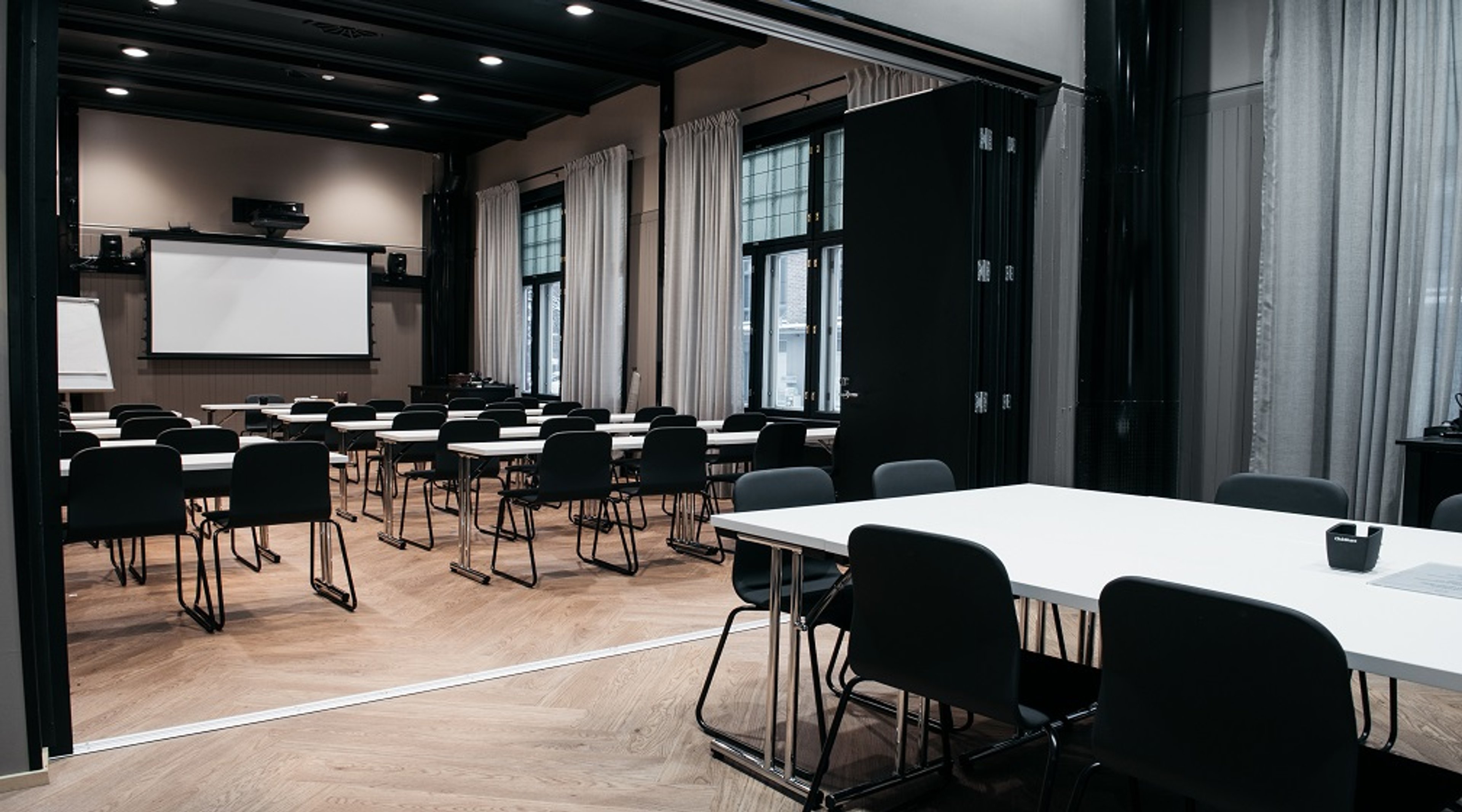 Modern conference room with projection screen, tables and chairs, light curtains covering large windows and wooden parquet flooring.
