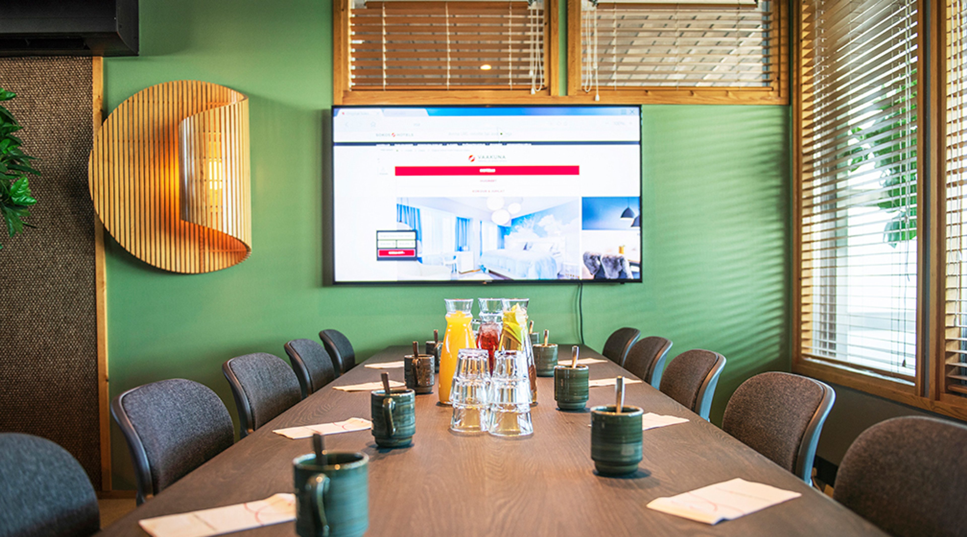Small meeting room with green walls and wooden table. Table has juice carafes and glasses. A large screen mounted on the wall.