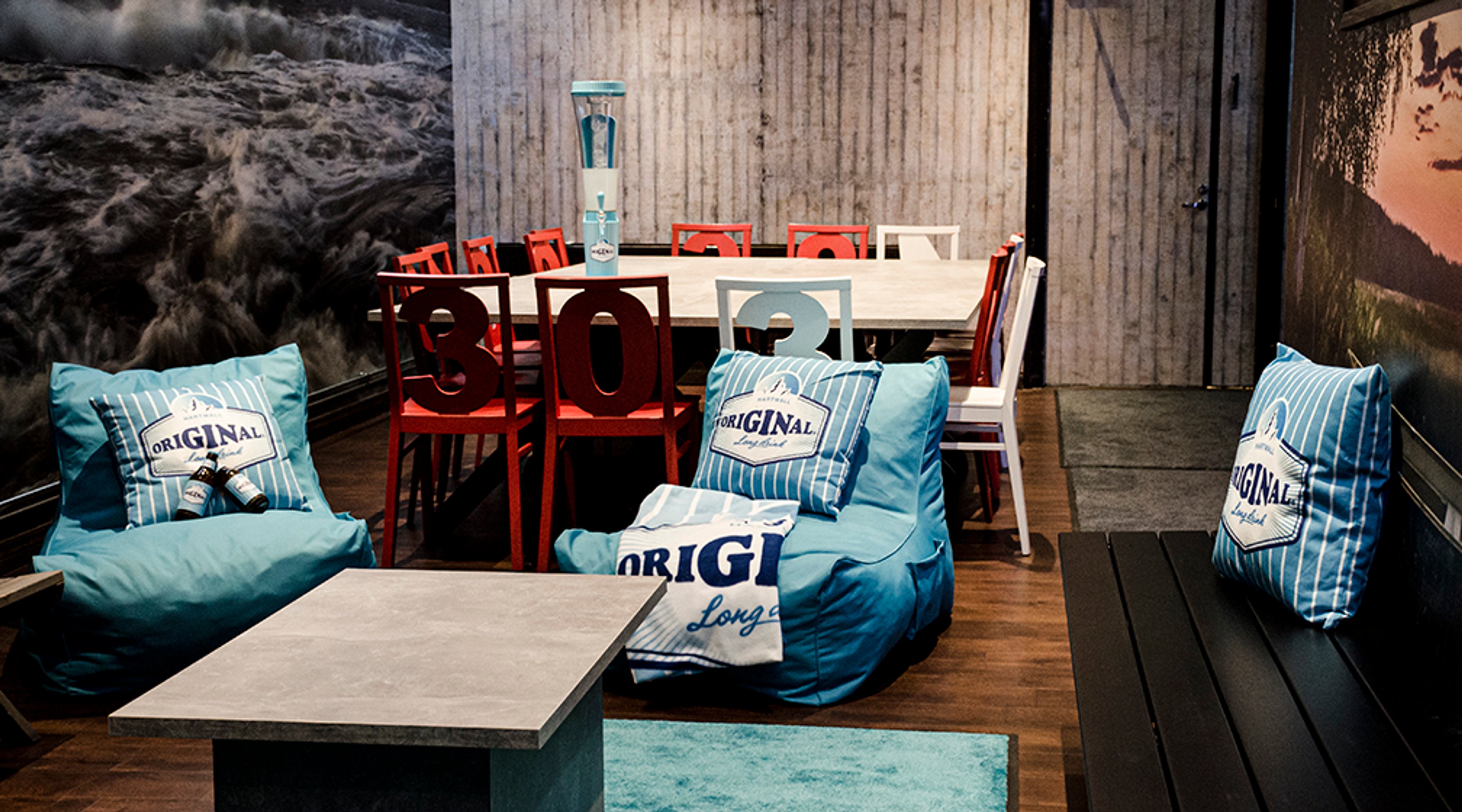 Lounge area with blue beanbag chairs, red and white chairs around a wooden table, sea-themed wall image and concrete wall in background.