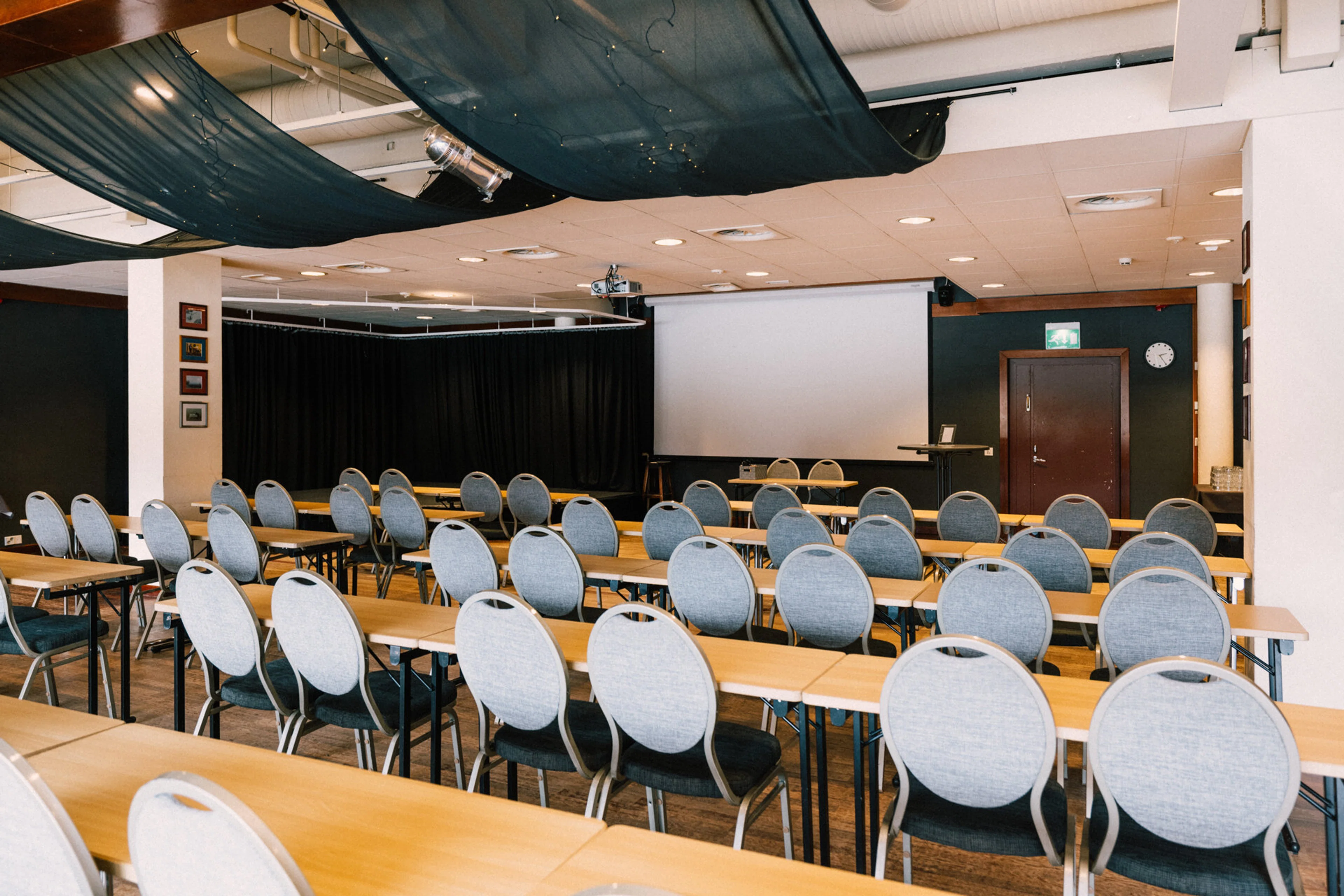 Konferensrum med bordsrader och stolar, projektorduk framme och svart gardin på sidan. Jämn belysning och brunt golv.
