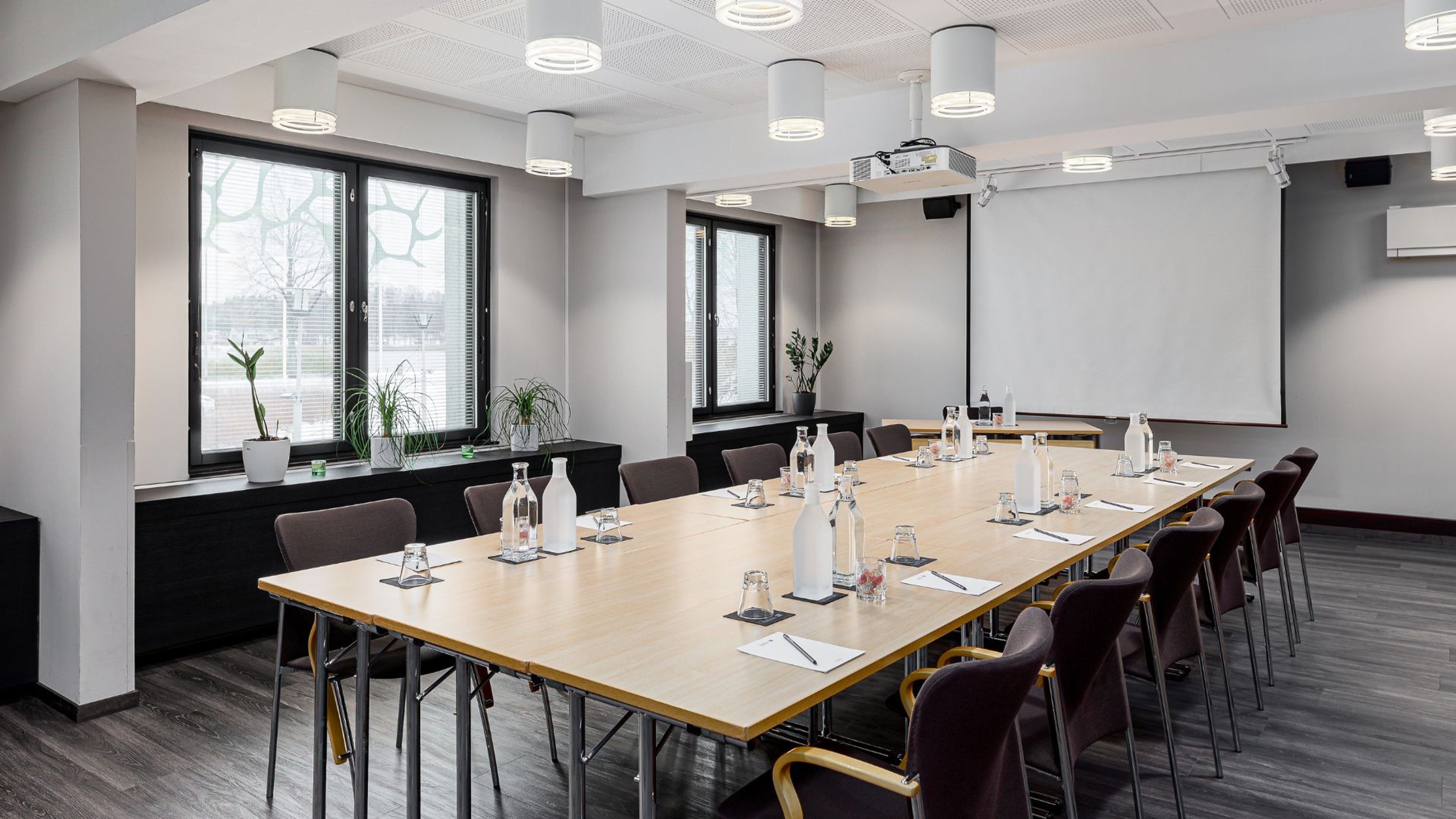 Moderni kokoushuone pitkällä puupöydällä, vesikarahveilla ja lasipulloilla. Tila on valoisa ikkunoiden, tasaisen valaistuksen ja projektorin ansiosta.