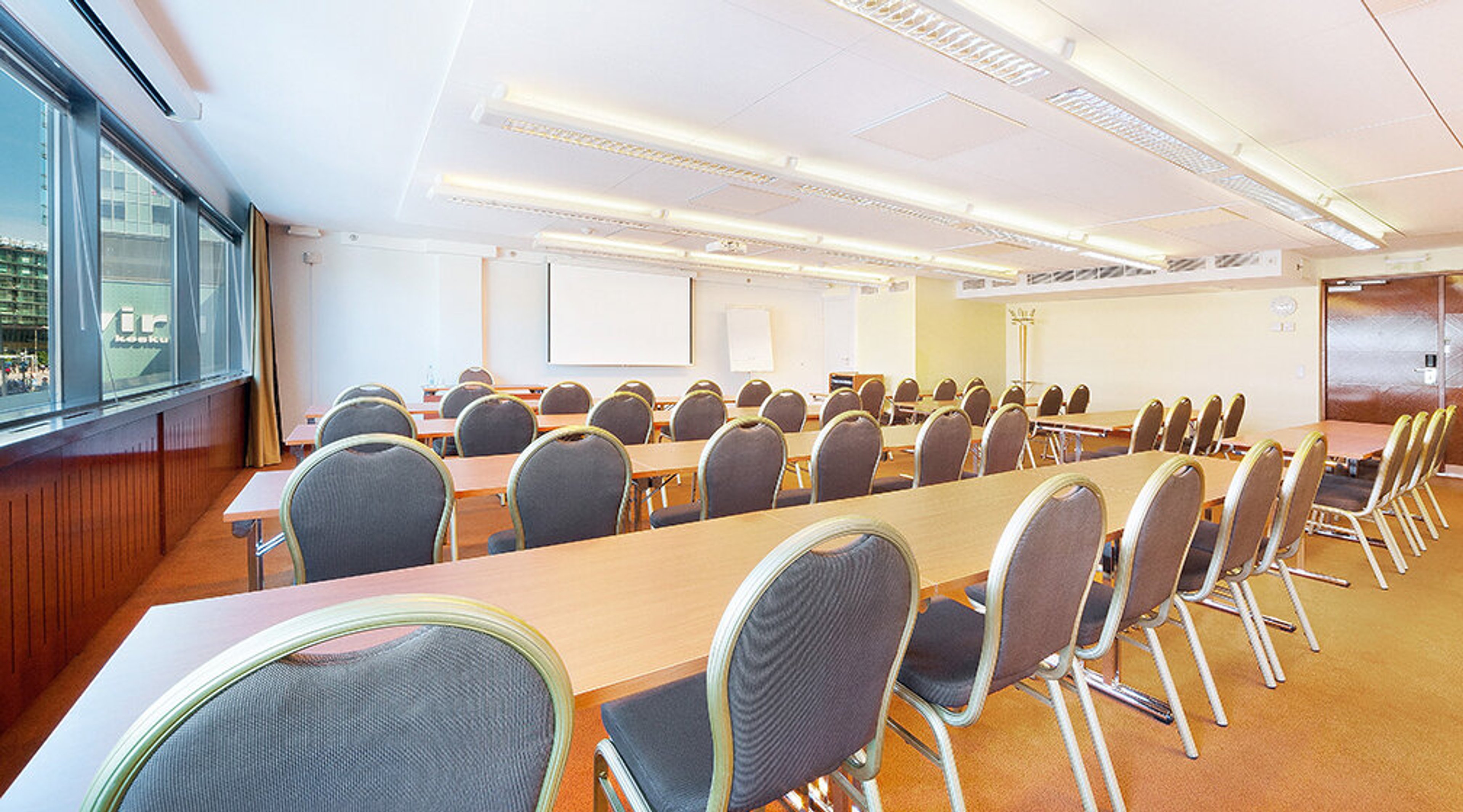 Bright conference room with long tables and chairs arranged in classroom style. White projector screen at the front and windows along the left wall.