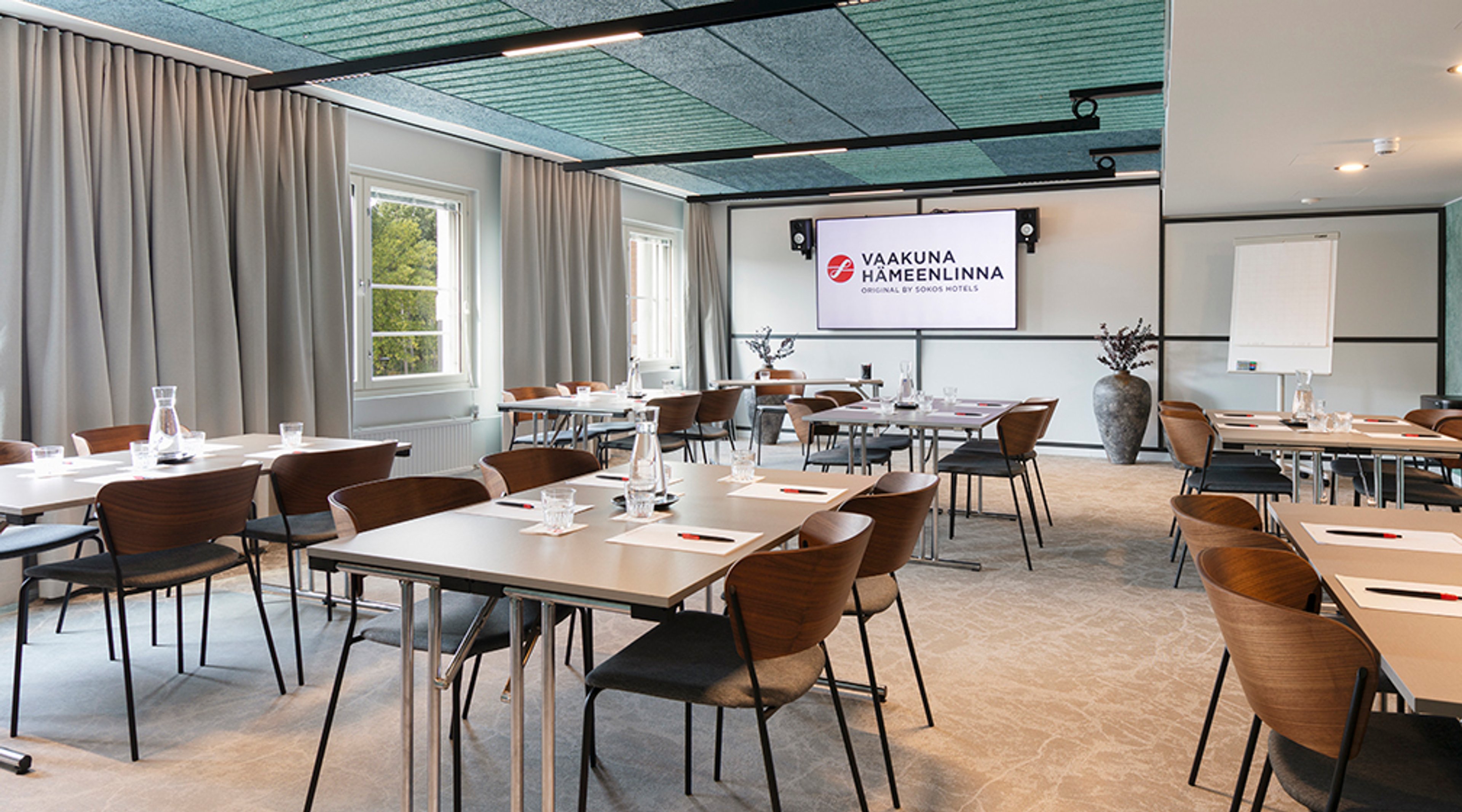 Modern meeting room with multiple tables and chairs, large windows, light carpet, and a presentation screen on the wall.