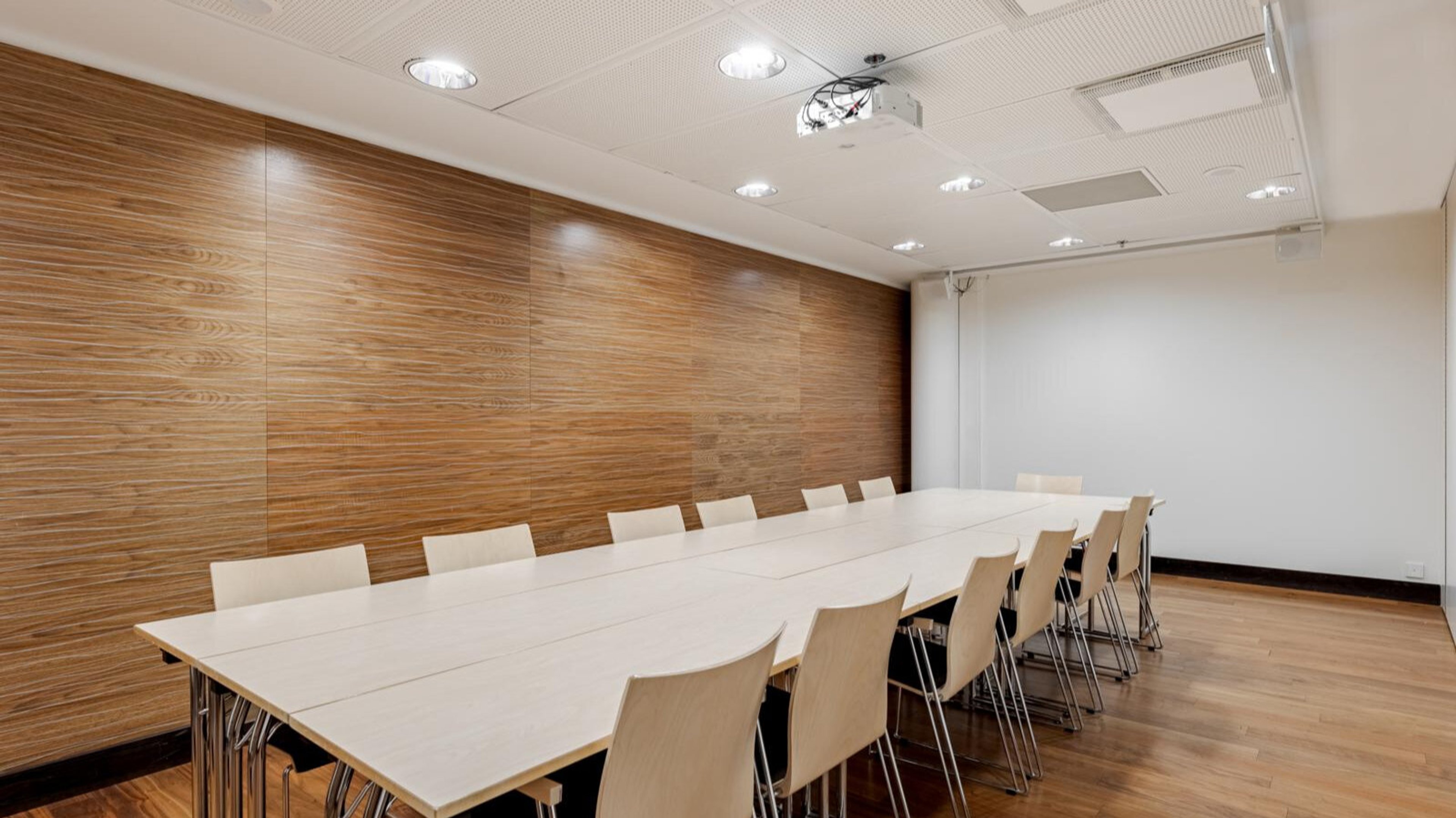 Modern conference room with long white table, light-colored chairs and wood-paneled wall. Space has even wooden flooring and ceiling-mounted projector.