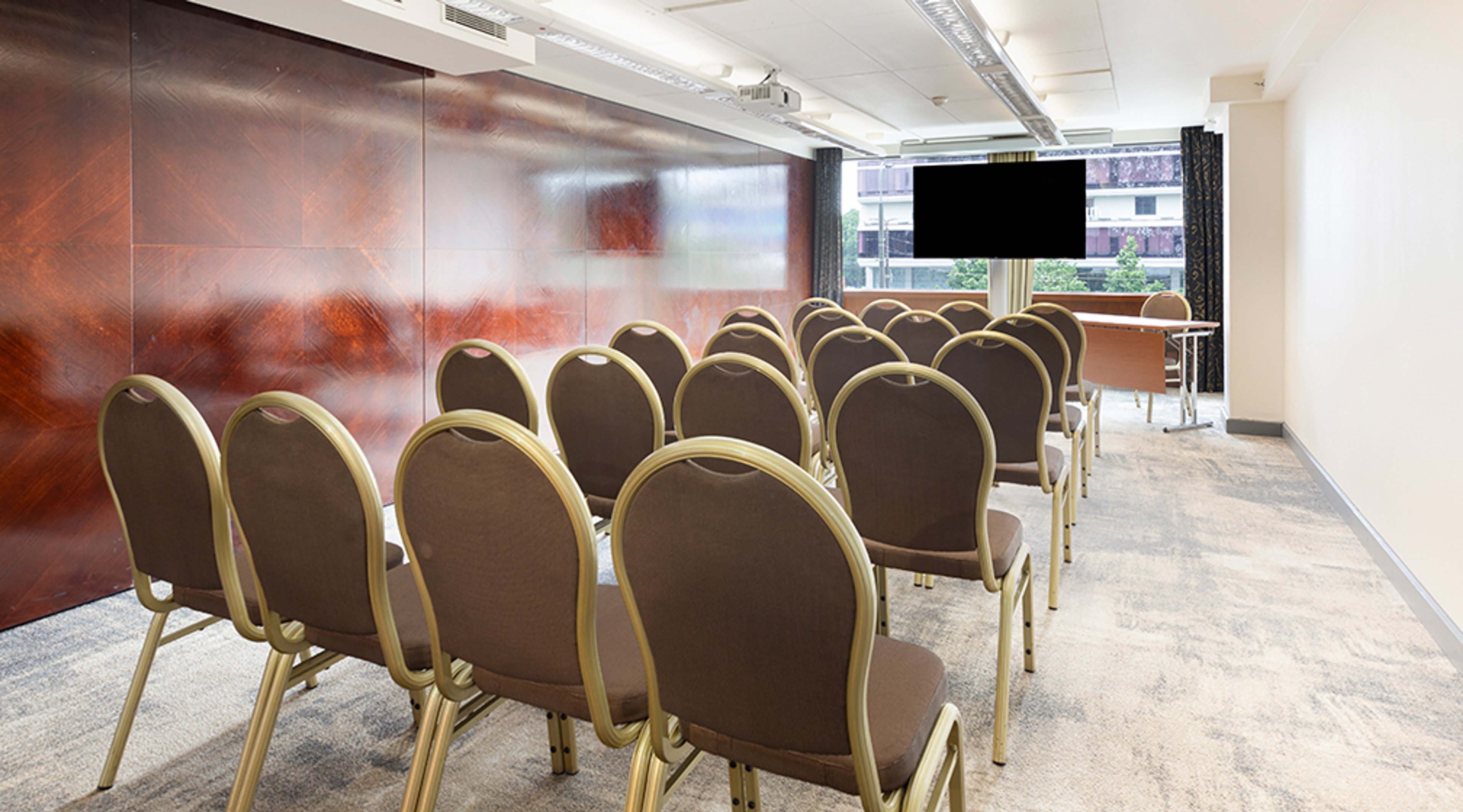 Meeting room with rows of chairs, wall-mounted television, and bright LED lighting. Space has hard flooring and wide aisles between rows.