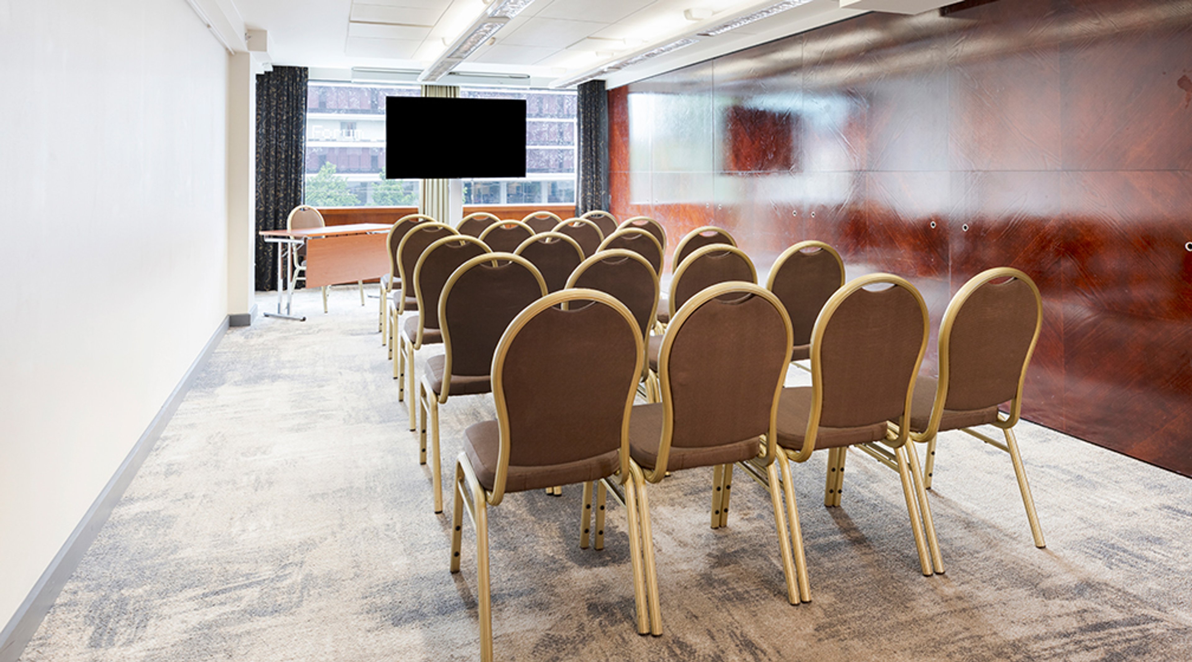 Meeting room with rows of brown upholstered chairs, large screen, glass wall and low-pile carpet flooring.