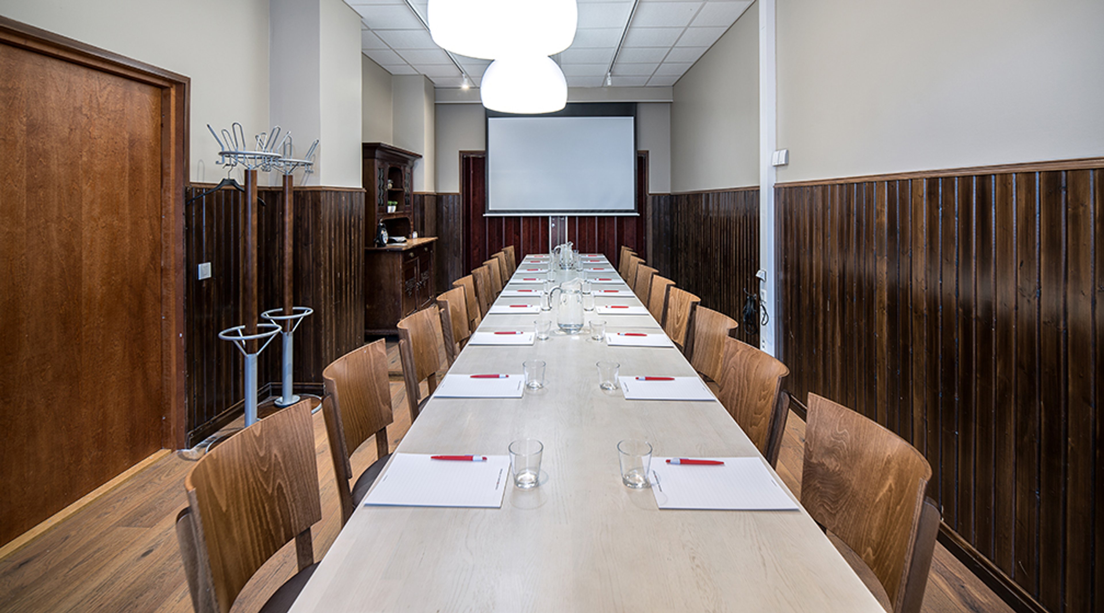 Long meeting room with wooden paneling. White table, wooden chairs, projector screen, glasses and notepads arranged on the table. High ceiling with lighting.