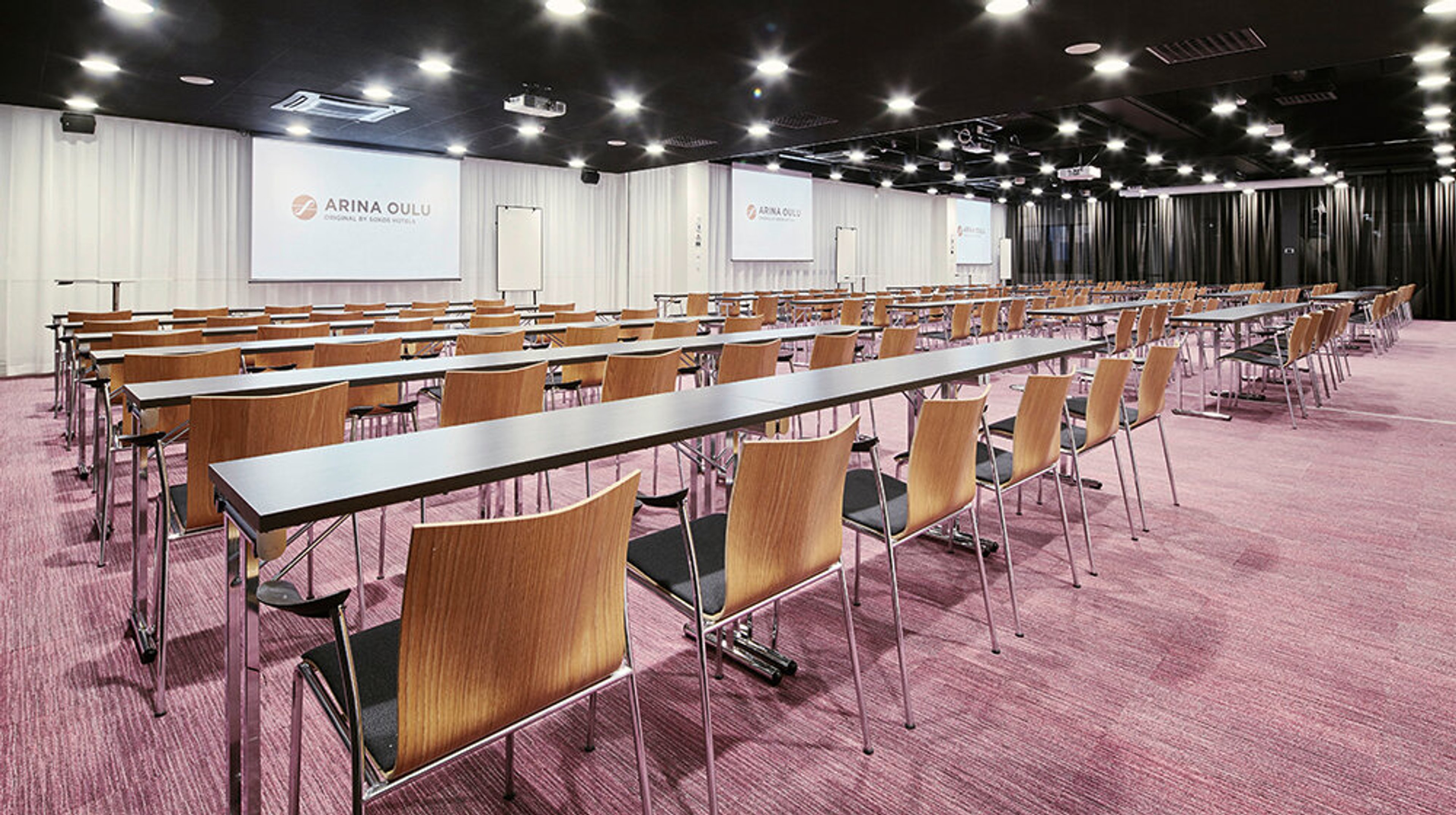 Conference room with long tables and wooden chairs arranged in rows. The space has pink wall-to-wall carpeting and black ceiling with bright lights.
