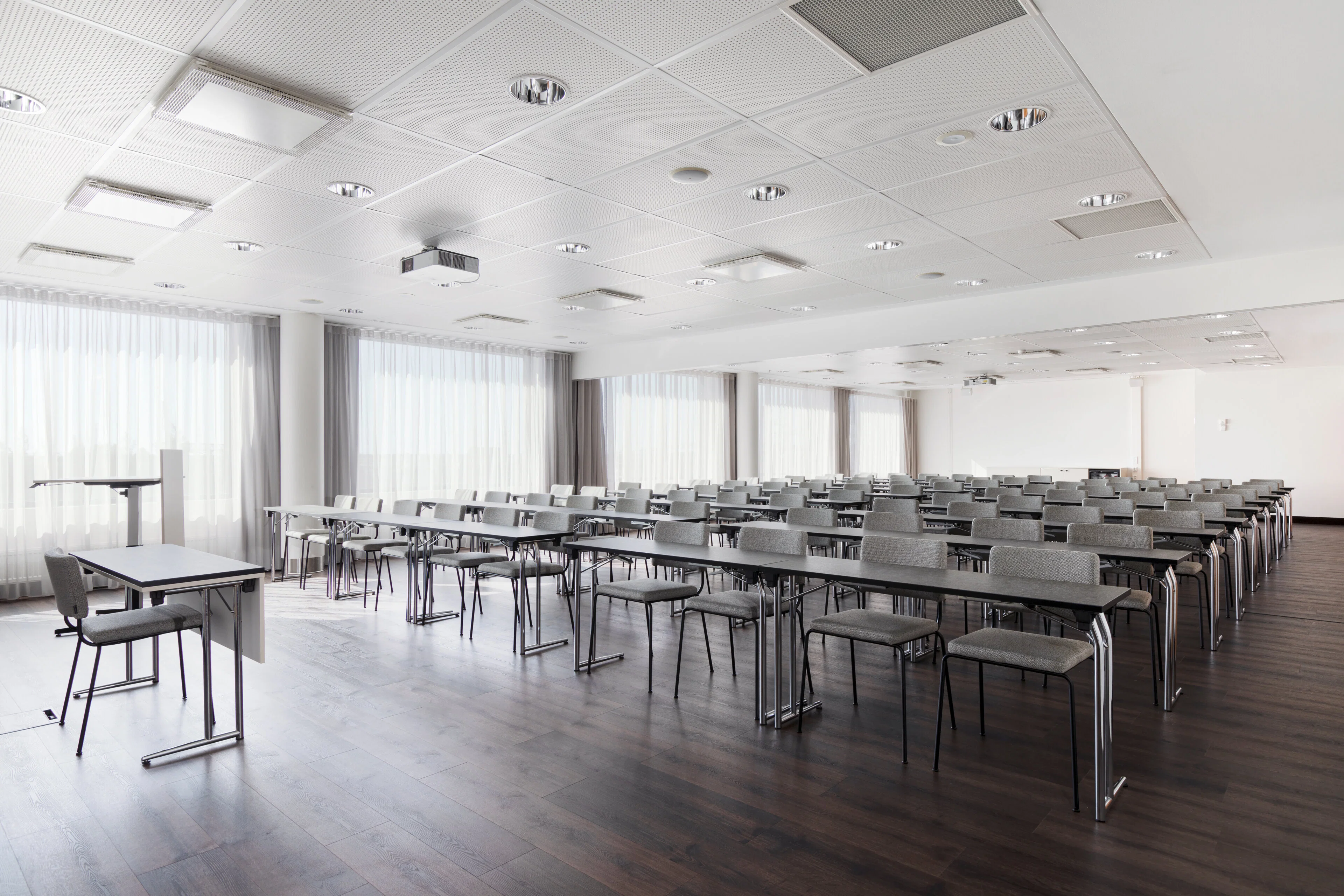 Modern conference room with long tables and gray chairs arranged in rows. The room features ceiling lights and white curtains on the walls.