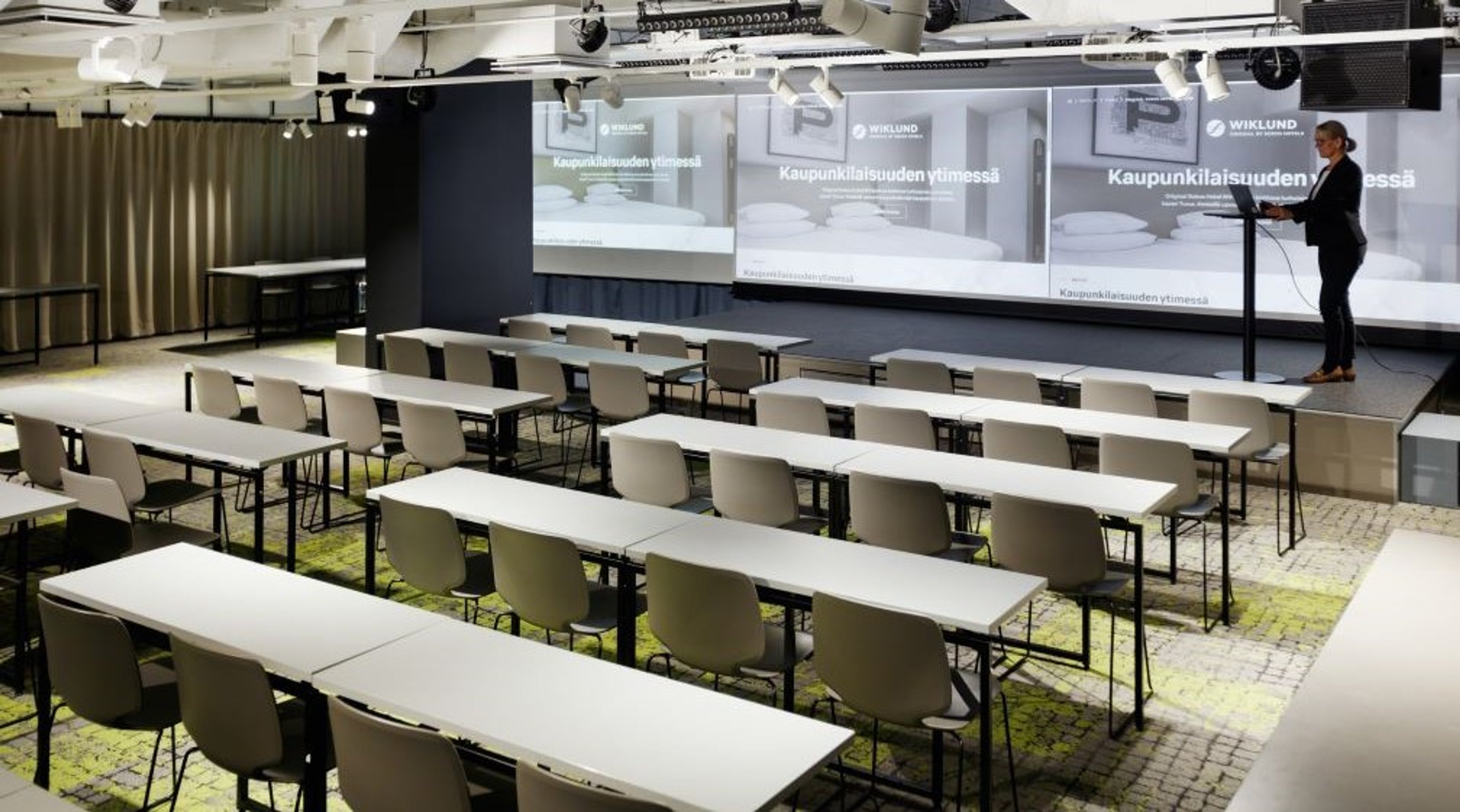 Modern classroom with long tables and white chairs. A presenter stands at a podium in front of large display screens. The space is spacious with good lighting.