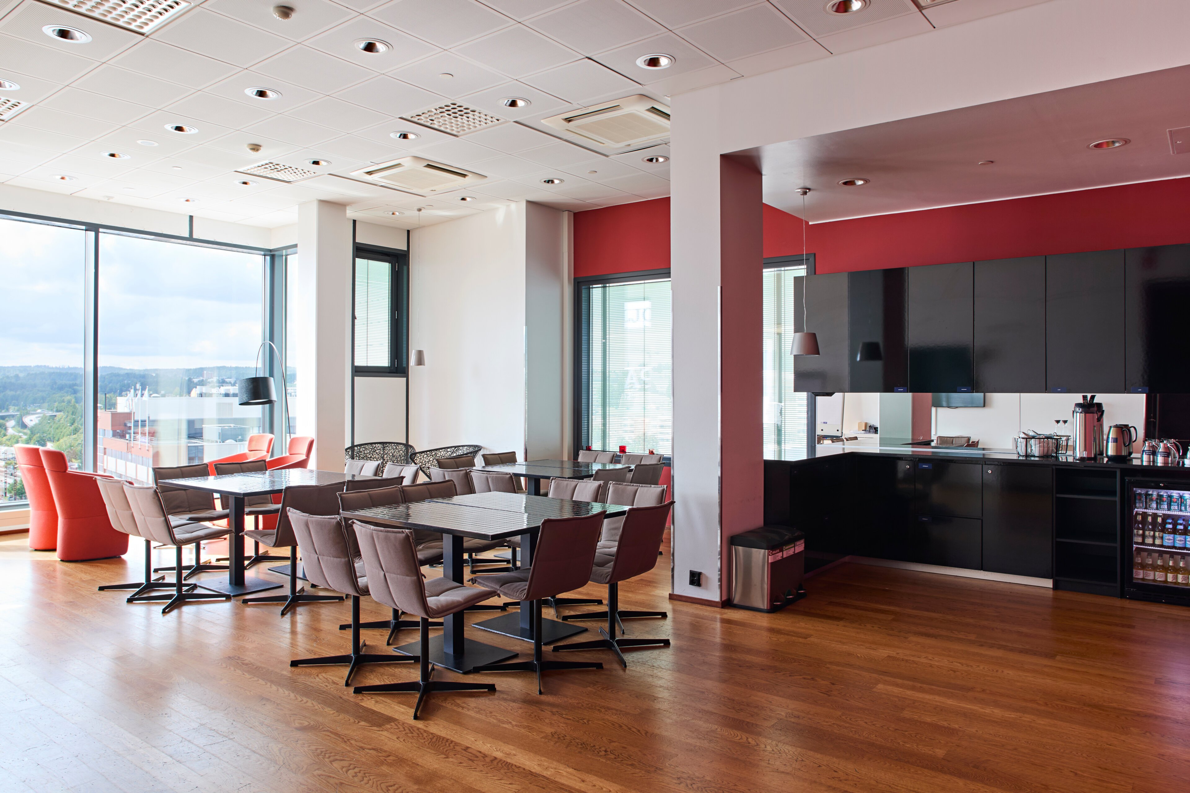 Modern break room with tables and chairs, open kitchen with black cabinets, red wall accents, and large windows overlooking city views.