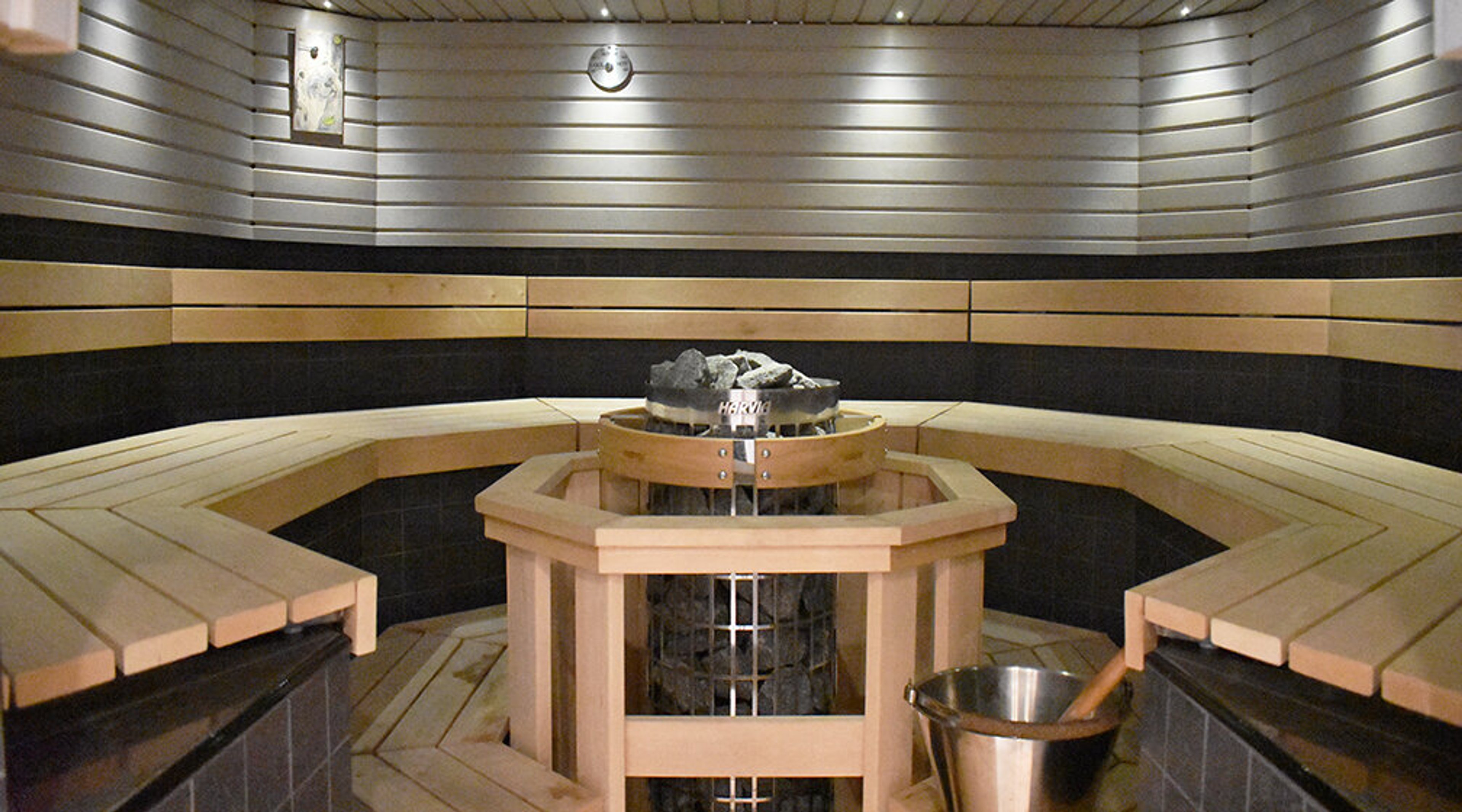 Modern sauna with wooden benches and hexagonal stove in the center. Light wooden panels adorn the wall surface.