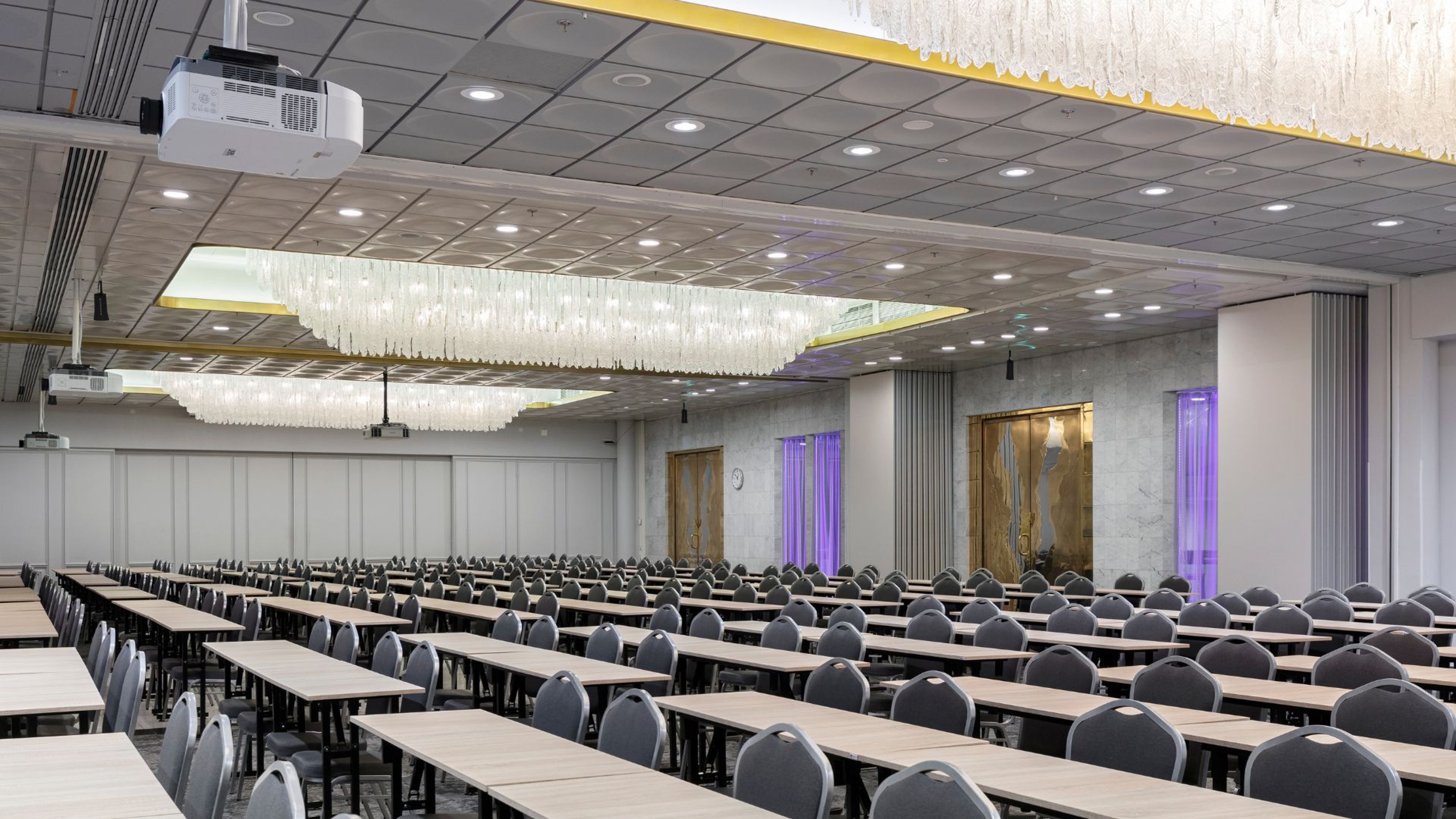 Suuri konferenssihuone rivistöillä pöytiä ja tuoleja, koristeelliset kristallivalaisimet katossa ja projektori valmiina esityksiä varten.