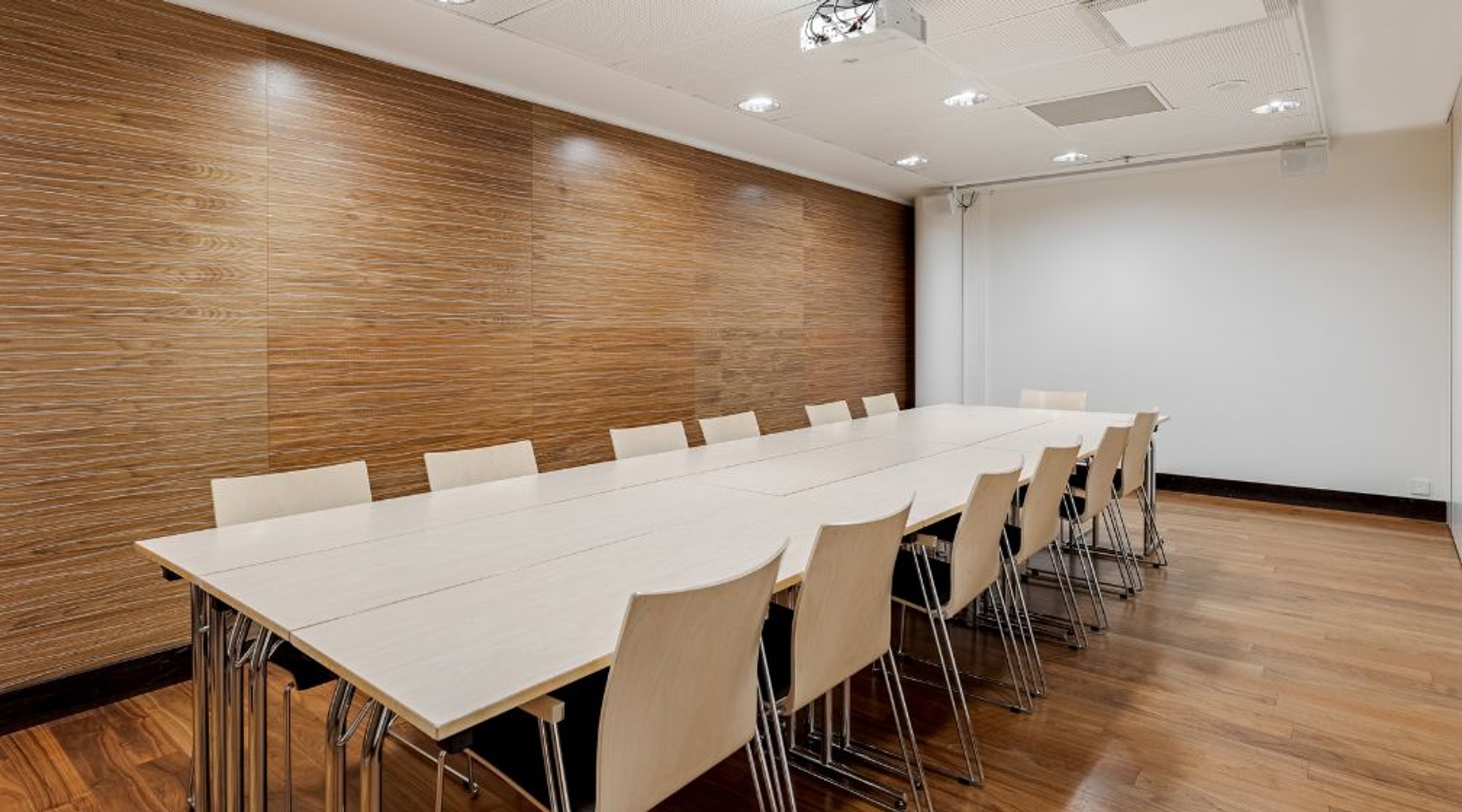 Modern conference room with a long white table, light-colored chairs and wooden panel wall. Room features wooden flooring and ceiling projector.