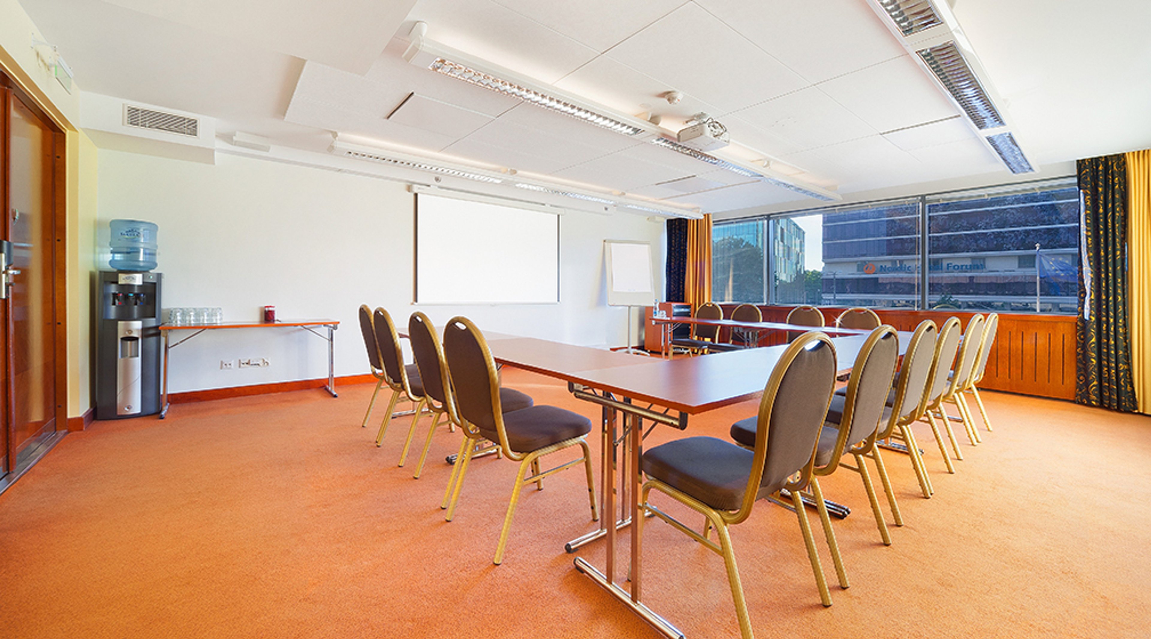 Meeting room with wooden table and chairs on orange carpet. Natural light from windows, water dispenser in corner.