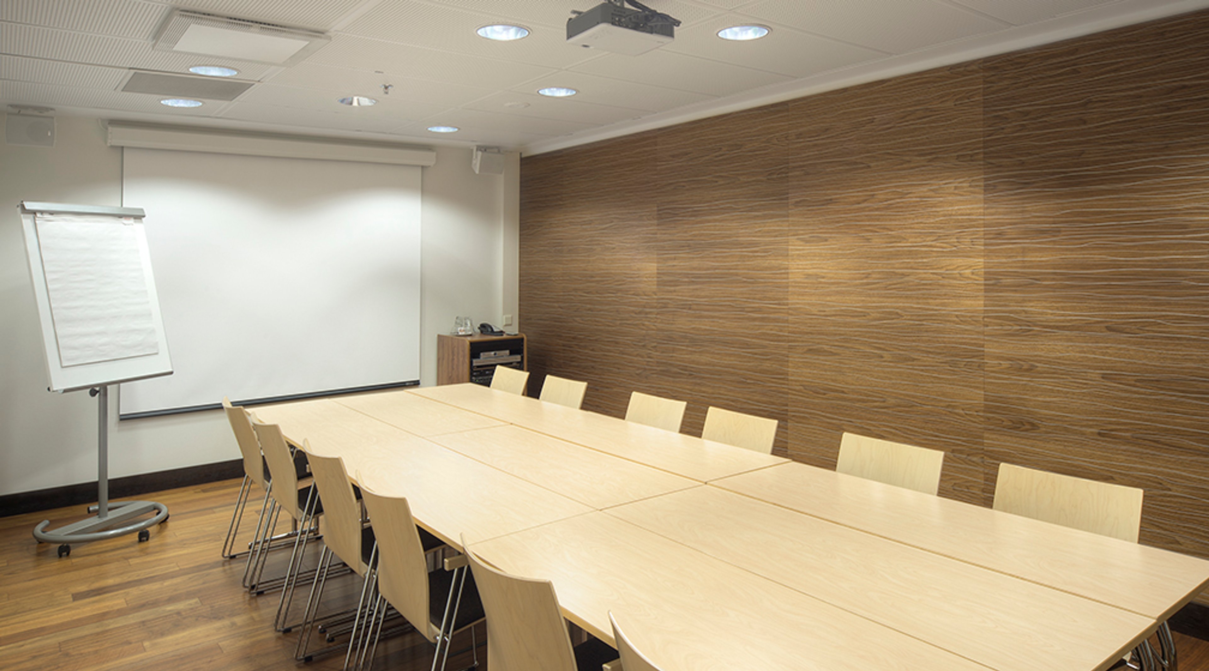 Modern meeting room with wooden panel wall, light-colored table, wooden chairs and a flip chart. Wooden floor, ceiling lights and projector.