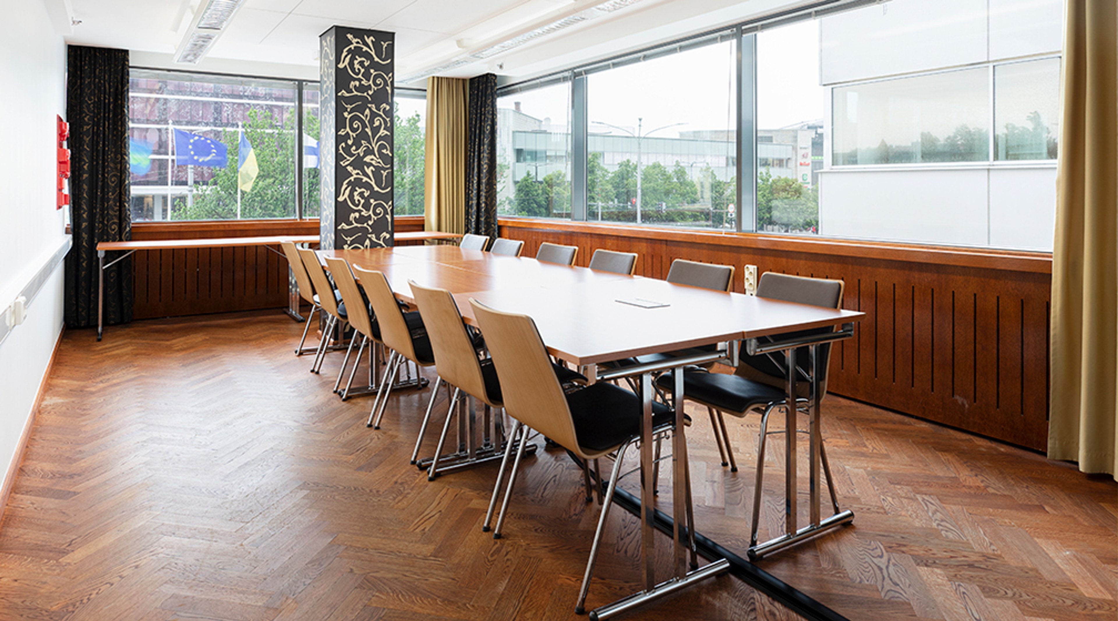 Meeting room with long table and chairs on parquet flooring, large windows and wooden wall panelling.