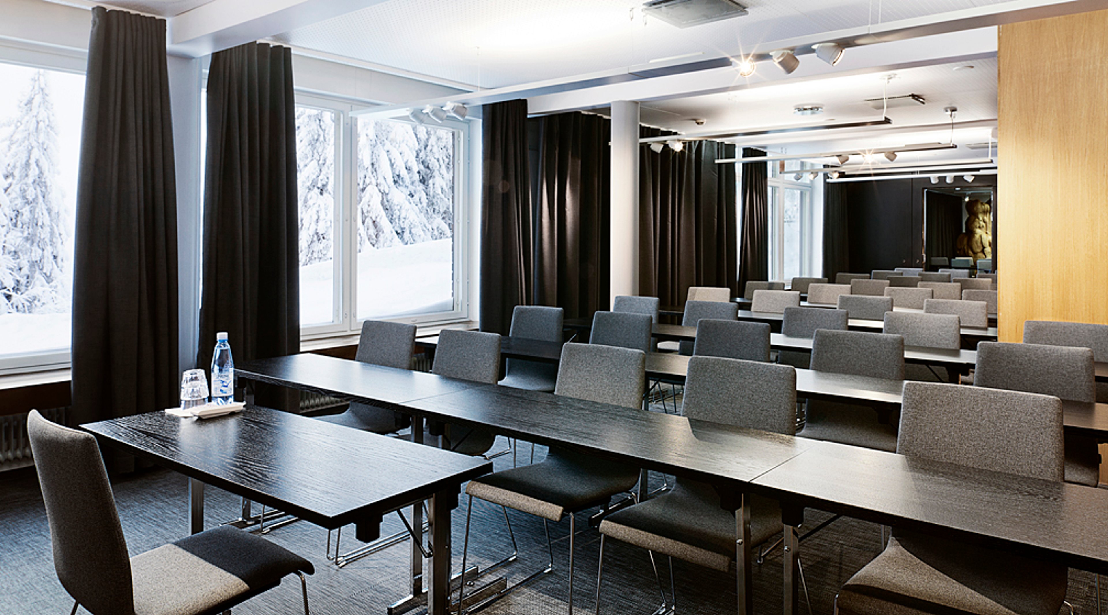 Modern auditorium med gråa bord och stolar, stora fönster visar snötäckta träd. Mörka gardiner och träpanel på väggen.