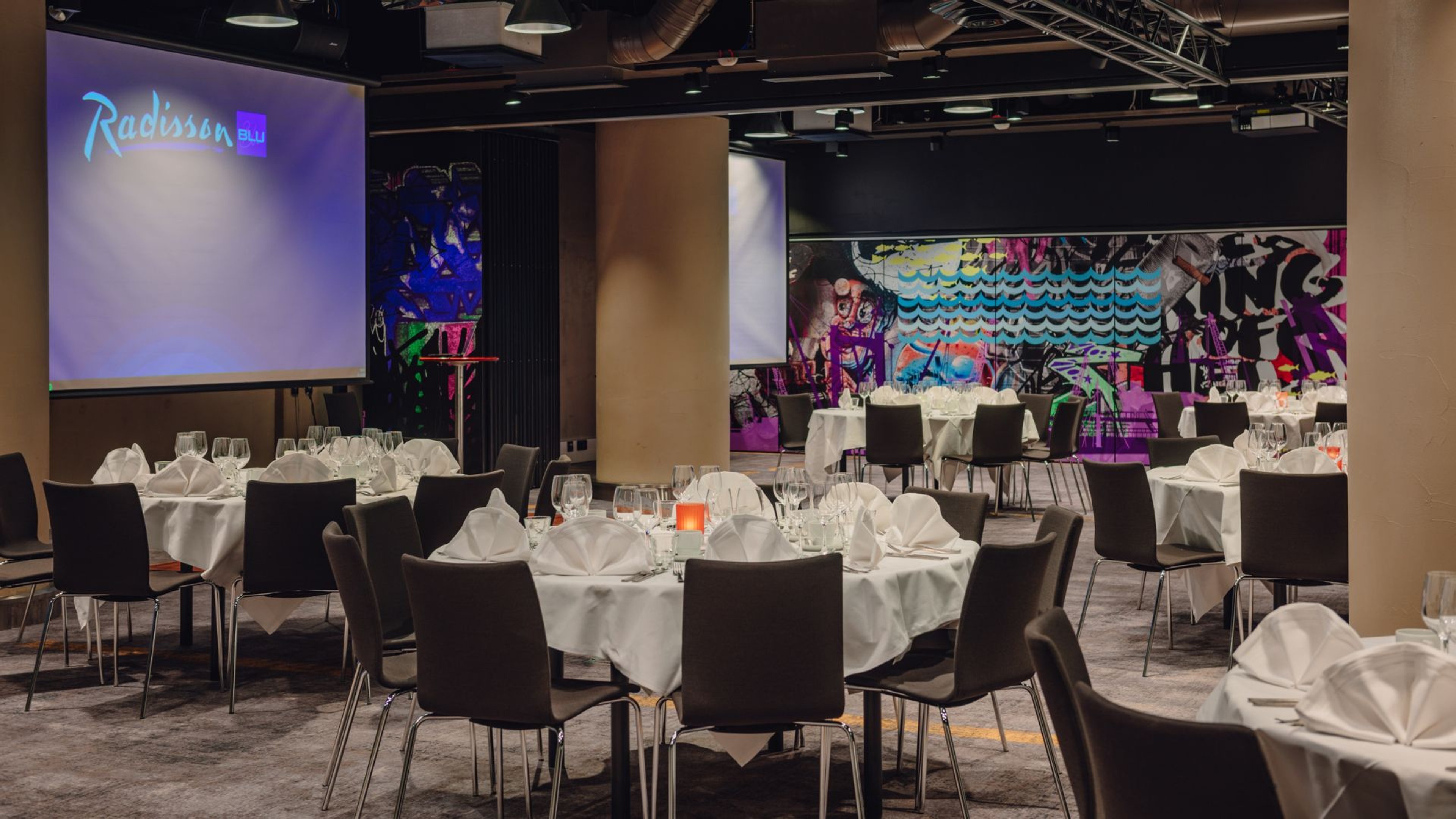 Radisson Blu -hotellin juhlatila, jossa on katettuja pyöreitä pöytiä, valkoinen pöytäliina ja tummat tuolit. Taustalla kaksi valkokangasta ja graffititaide seinällä.