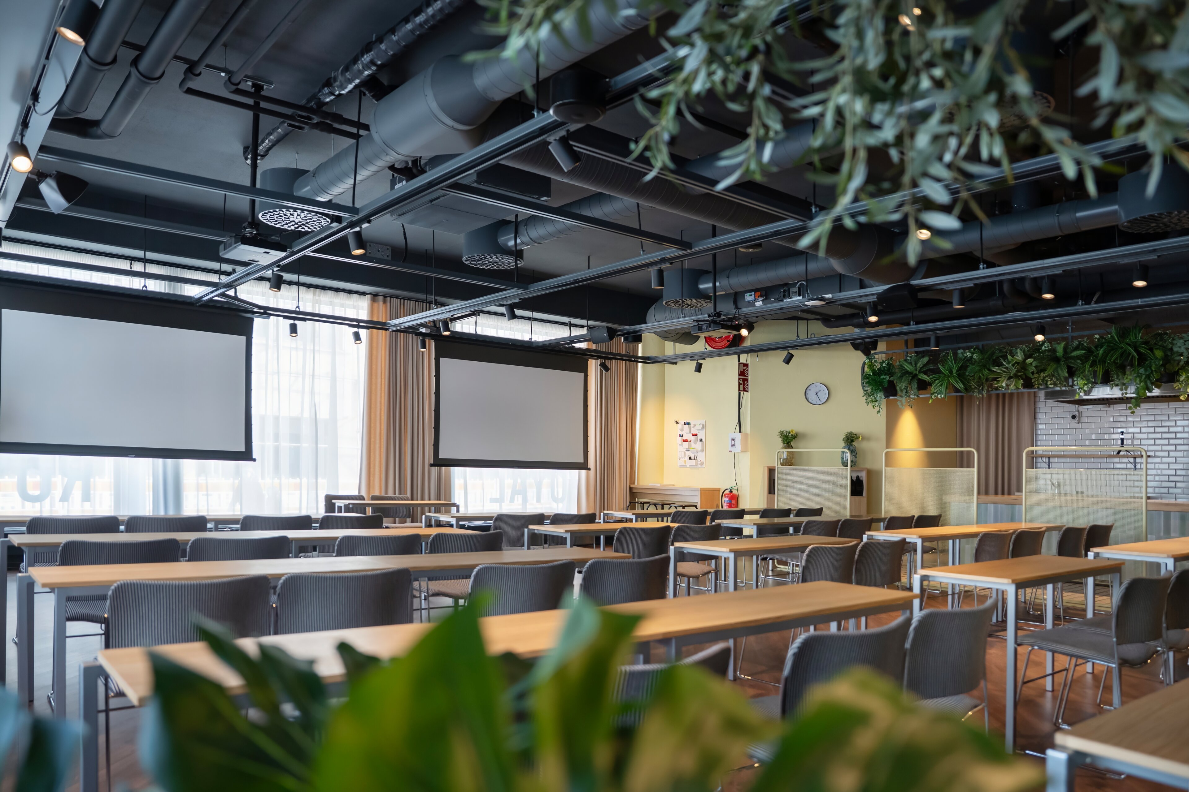 Modern meeting room with wooden tables and grey chairs. The space features two projector screens, indoor plants and industrial ceiling.