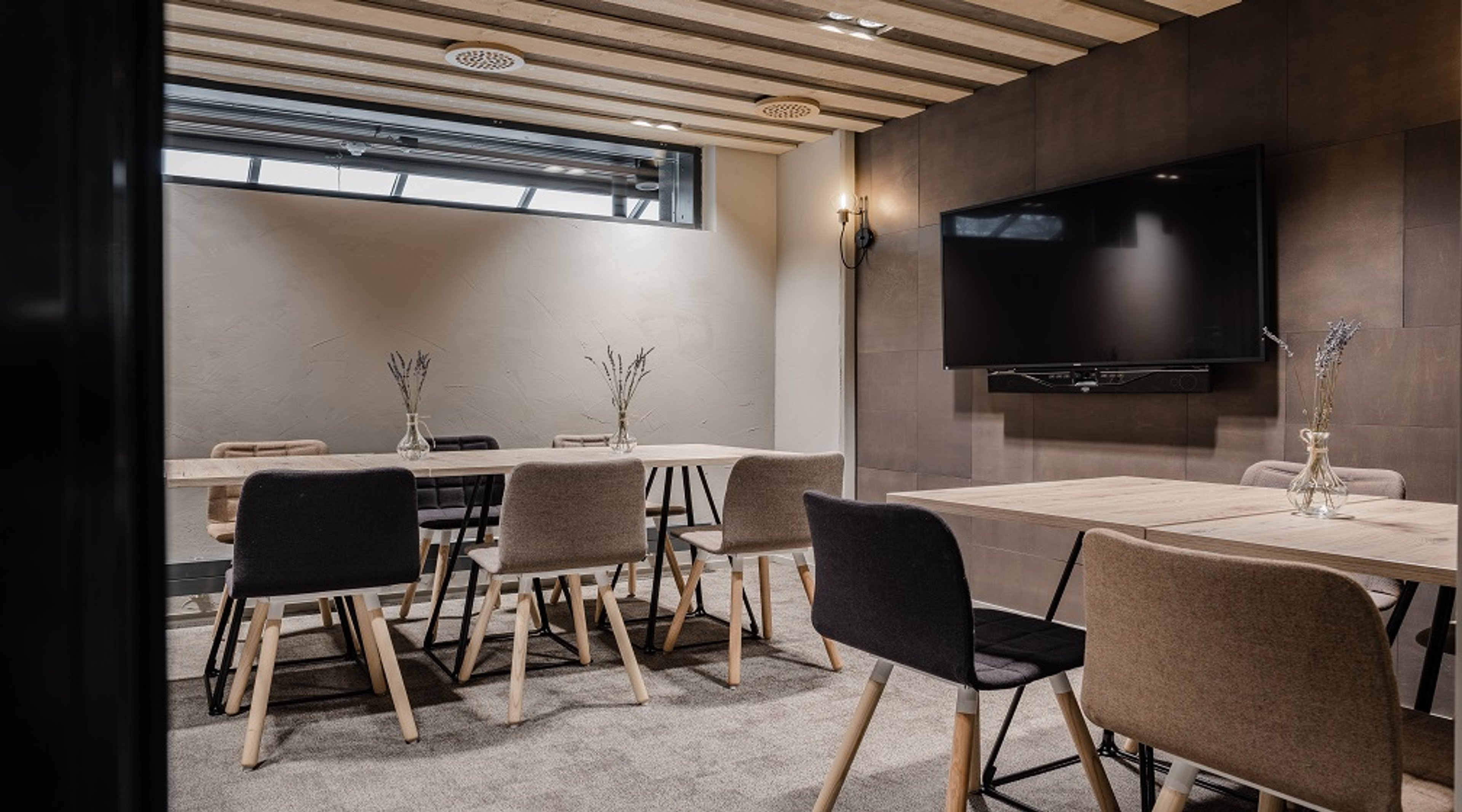 Modern meeting room with two wooden tables, chairs, wall-mounted TV, and high windows. Floor covered with soft carpeting.