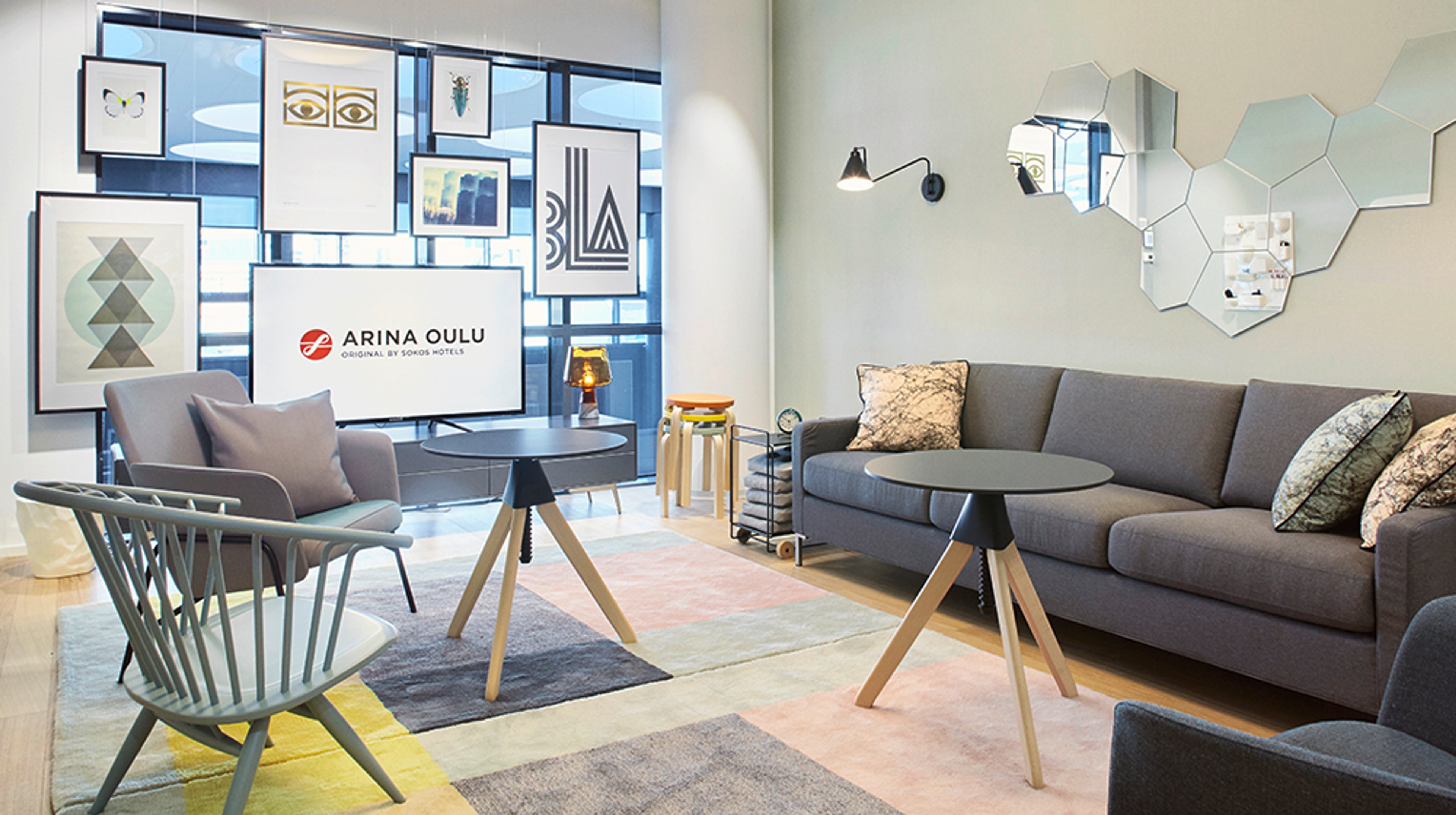 Stylish hotel lounge area with gray sofa, round tables and designer chairs. Patterned carpet and art on walls with a cluster of hexagonal mirrors.