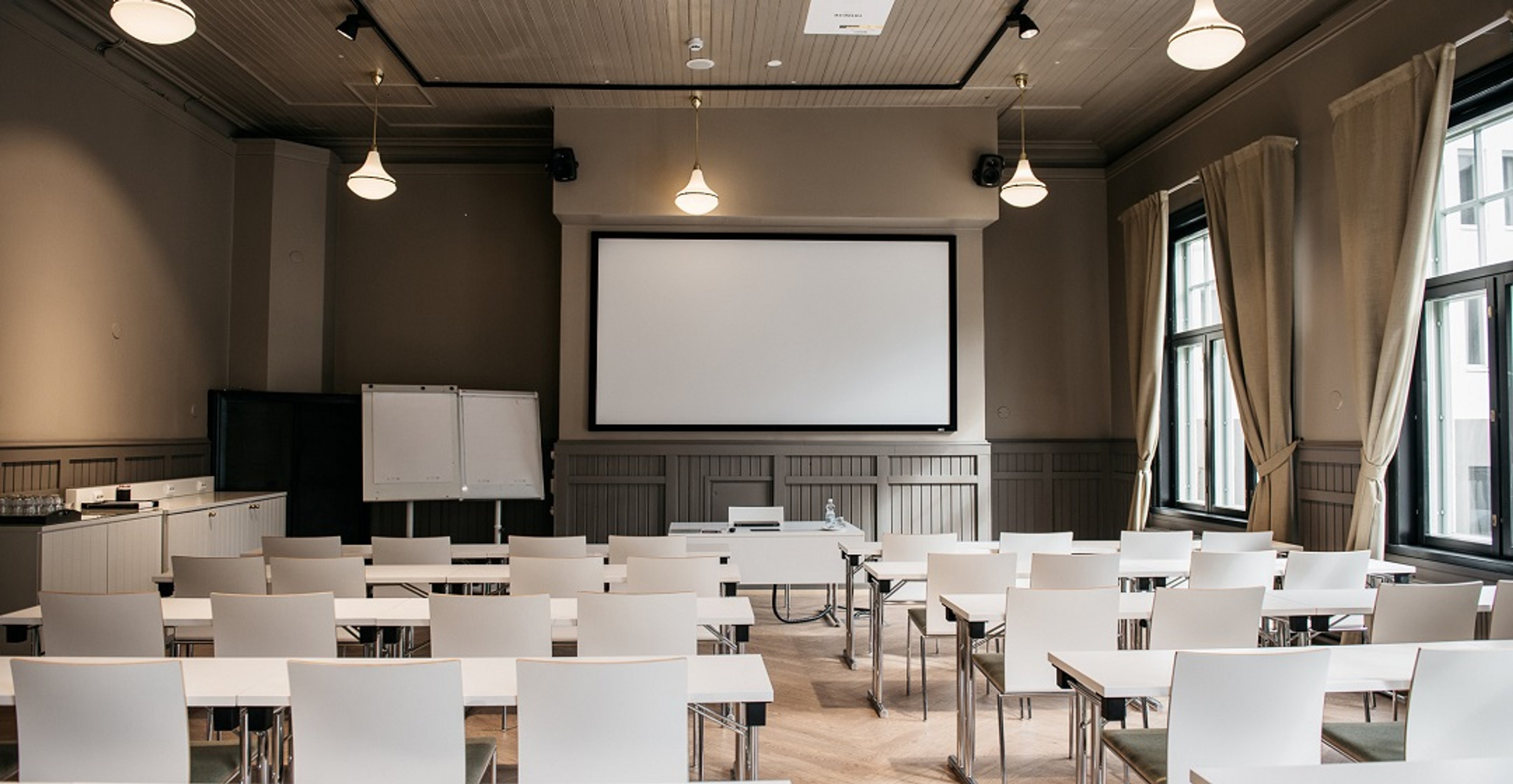 Conference room with white chairs at tables, projection screen at the front, large windows with beige curtains, and wooden floor.
