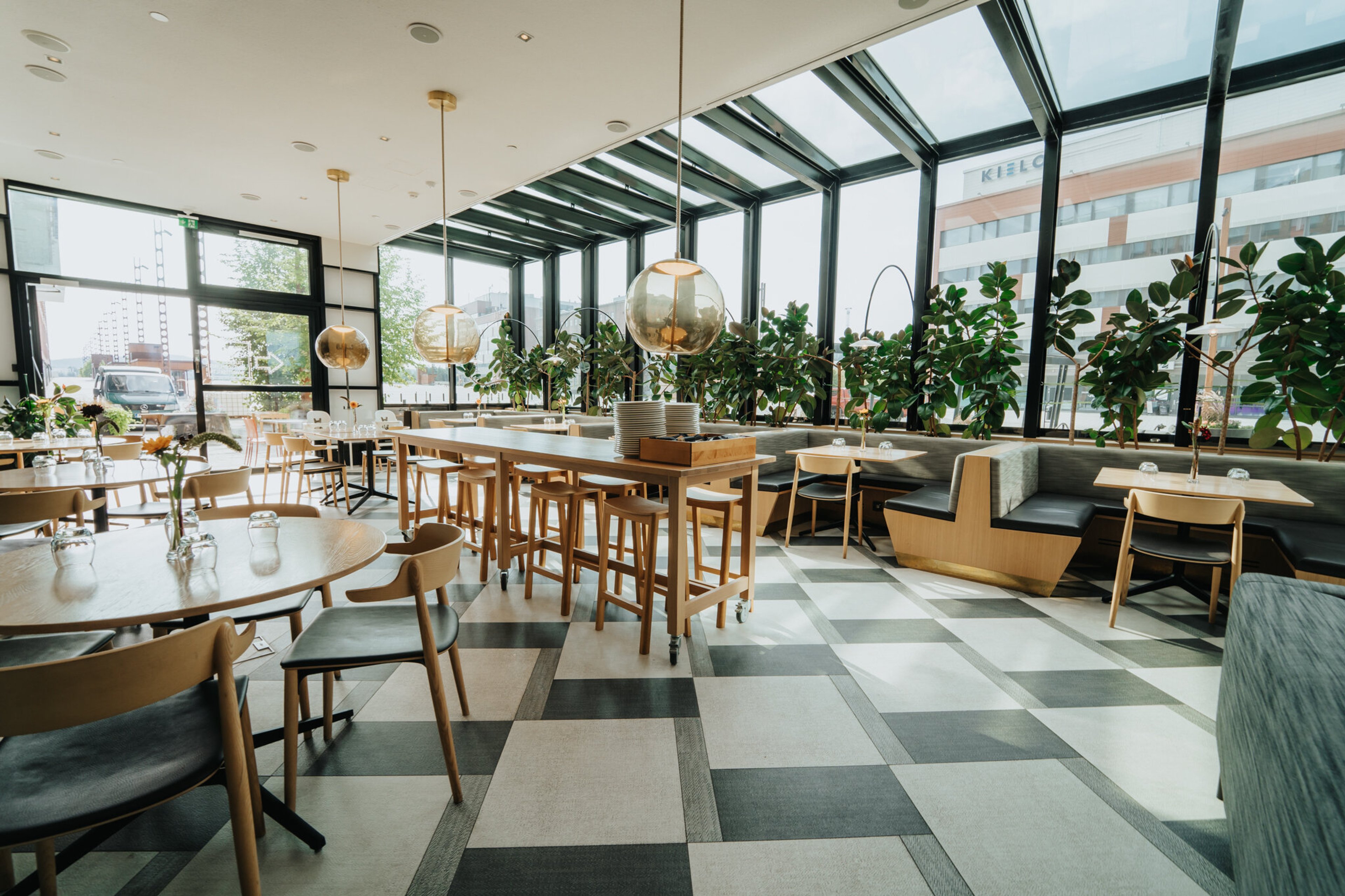 Bright restaurant with large windows, wooden tables and chairs, green plants and level flooring. Pendant lights hang from high ceiling.