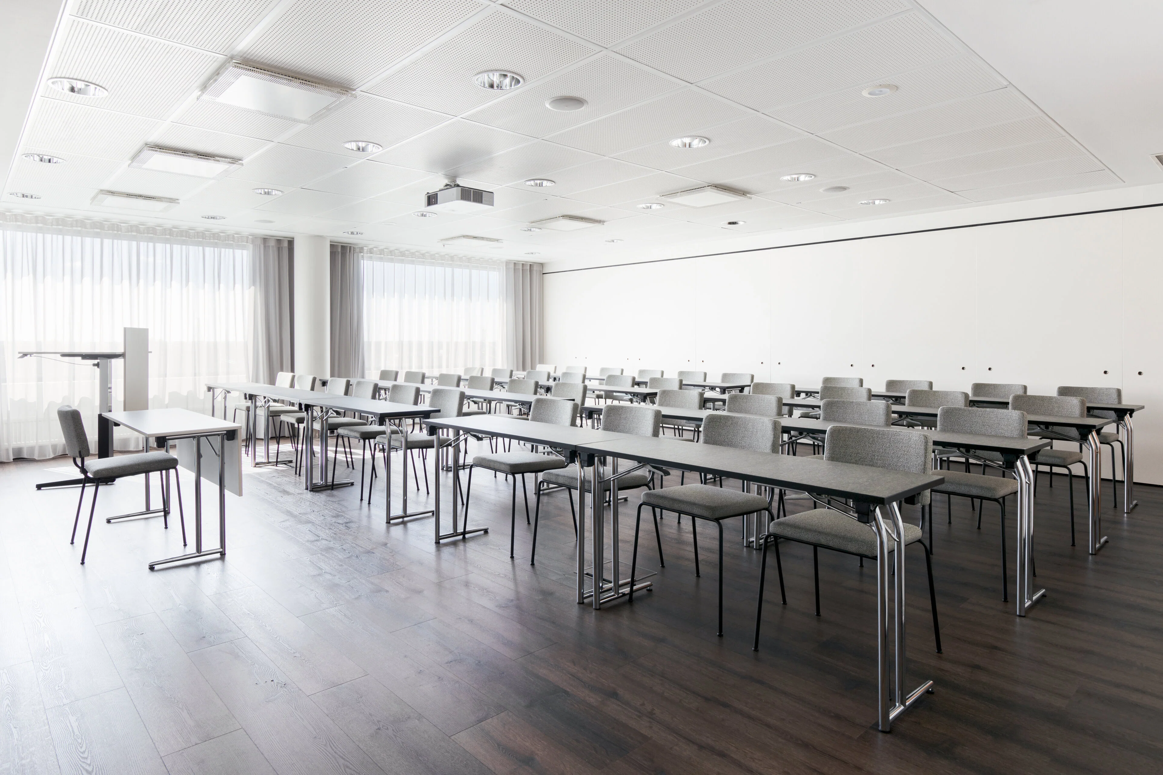 Modern, bright conference room with long tables and gray chairs arranged in rows. The room has a flat wooden floor and good ceiling lighting.