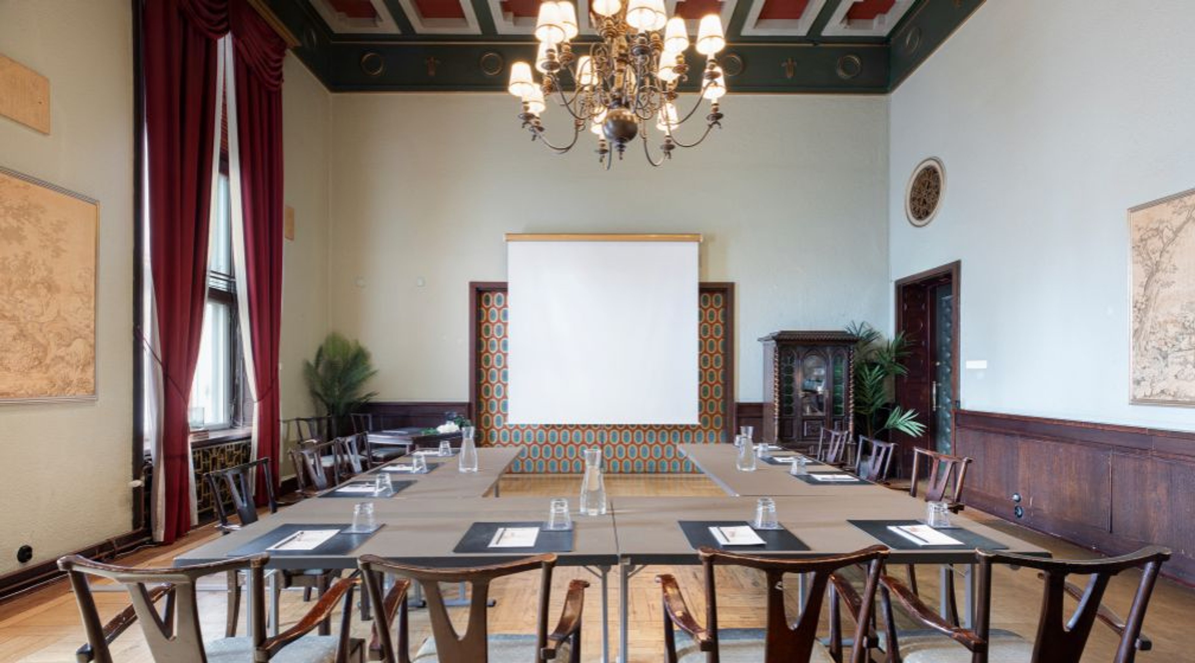 Conference room with long table and chairs, chandelier and wooden floor, projection screen in background.