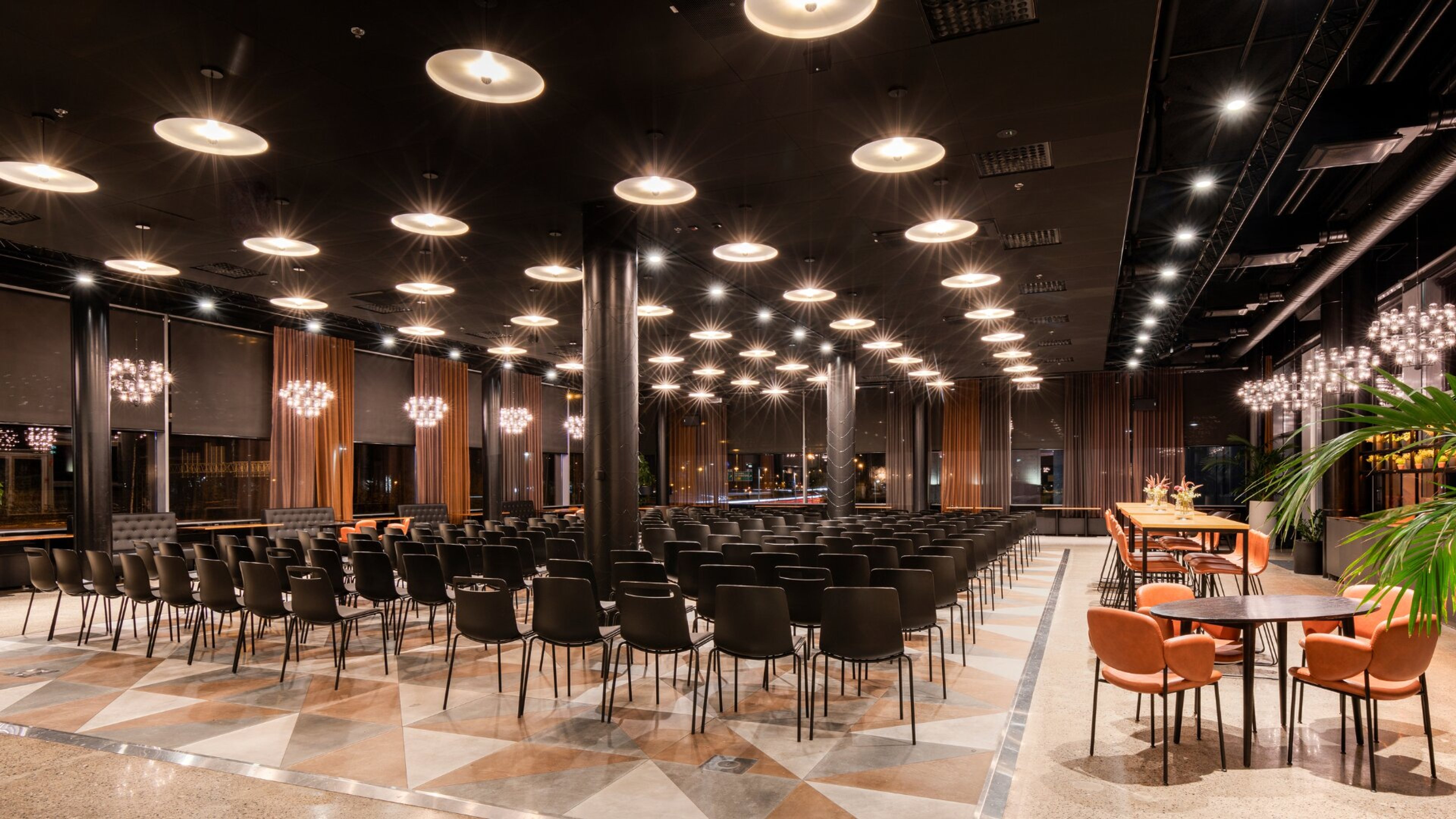 Modern auditorium with rows of black chairs, orange curtains, and striking ceiling lights. Floor features geometric patterns.