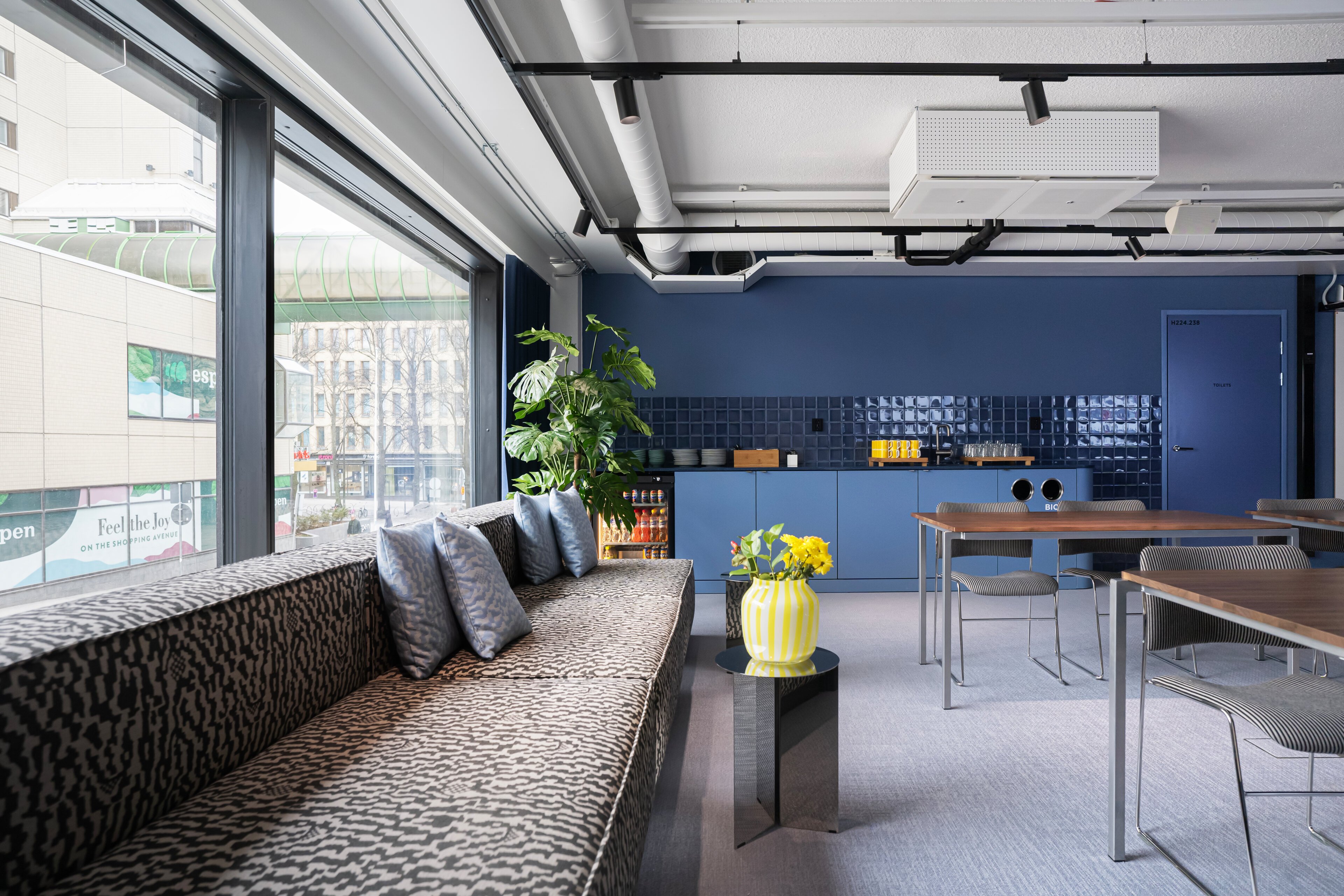 Modern blue common space with leopard-print sofa, tables, chairs, kitchenette and large window. The room features a yellow vase and green plants.