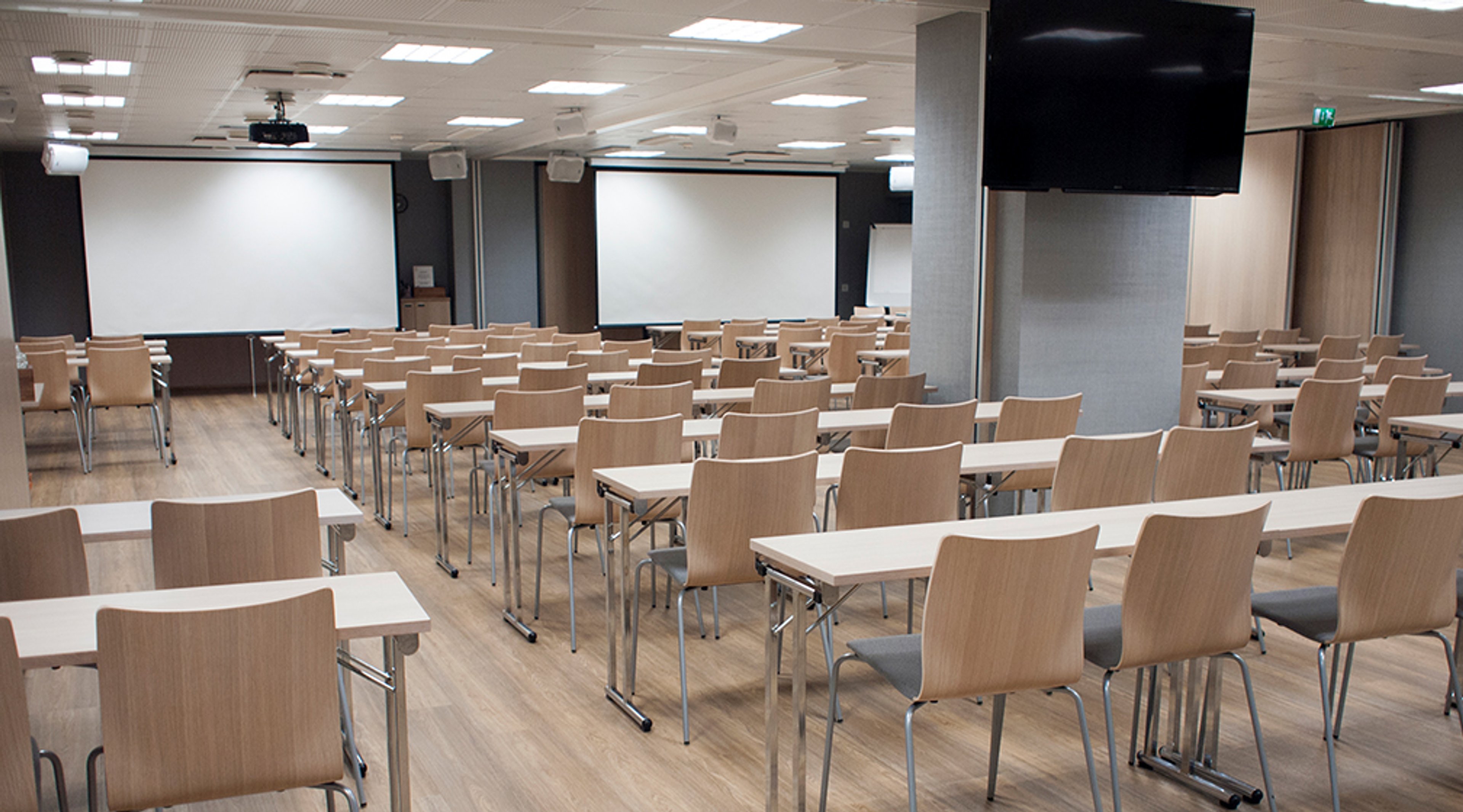 Modern auditorium with wooden chairs and tables, two white projector screens and large displays on walls, wooden flooring and bright lighting.