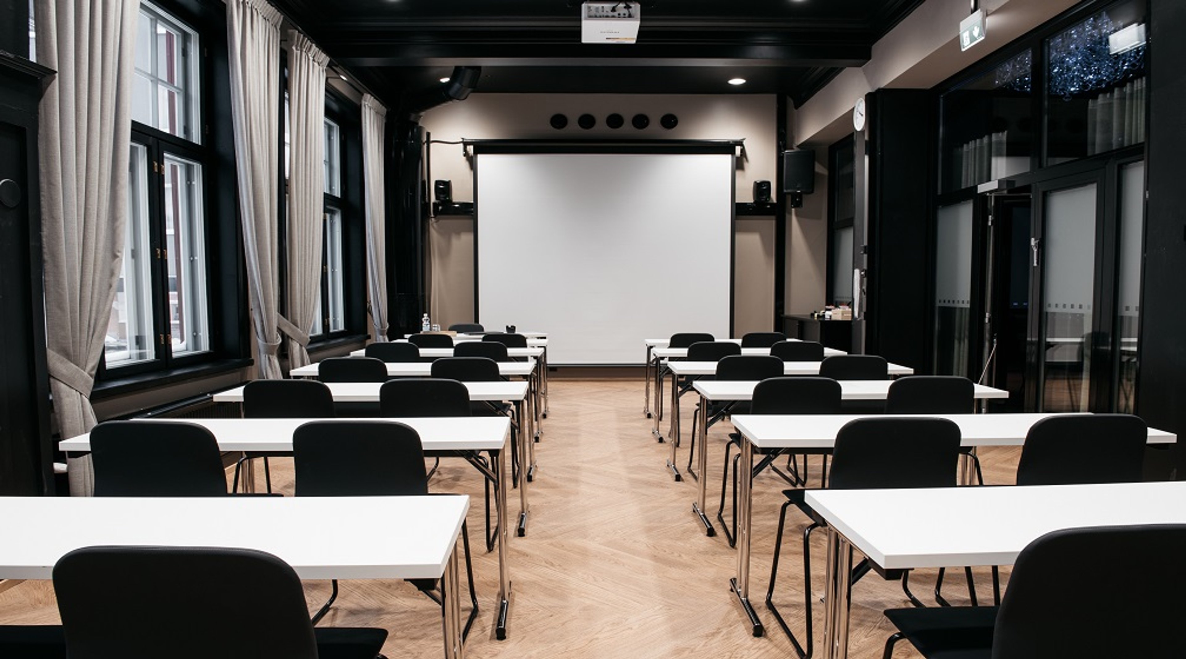 Conference room with white tables and black chairs arranged in rows, projection screen at front and large windows along the side. Wooden floor and black ceiling structures.