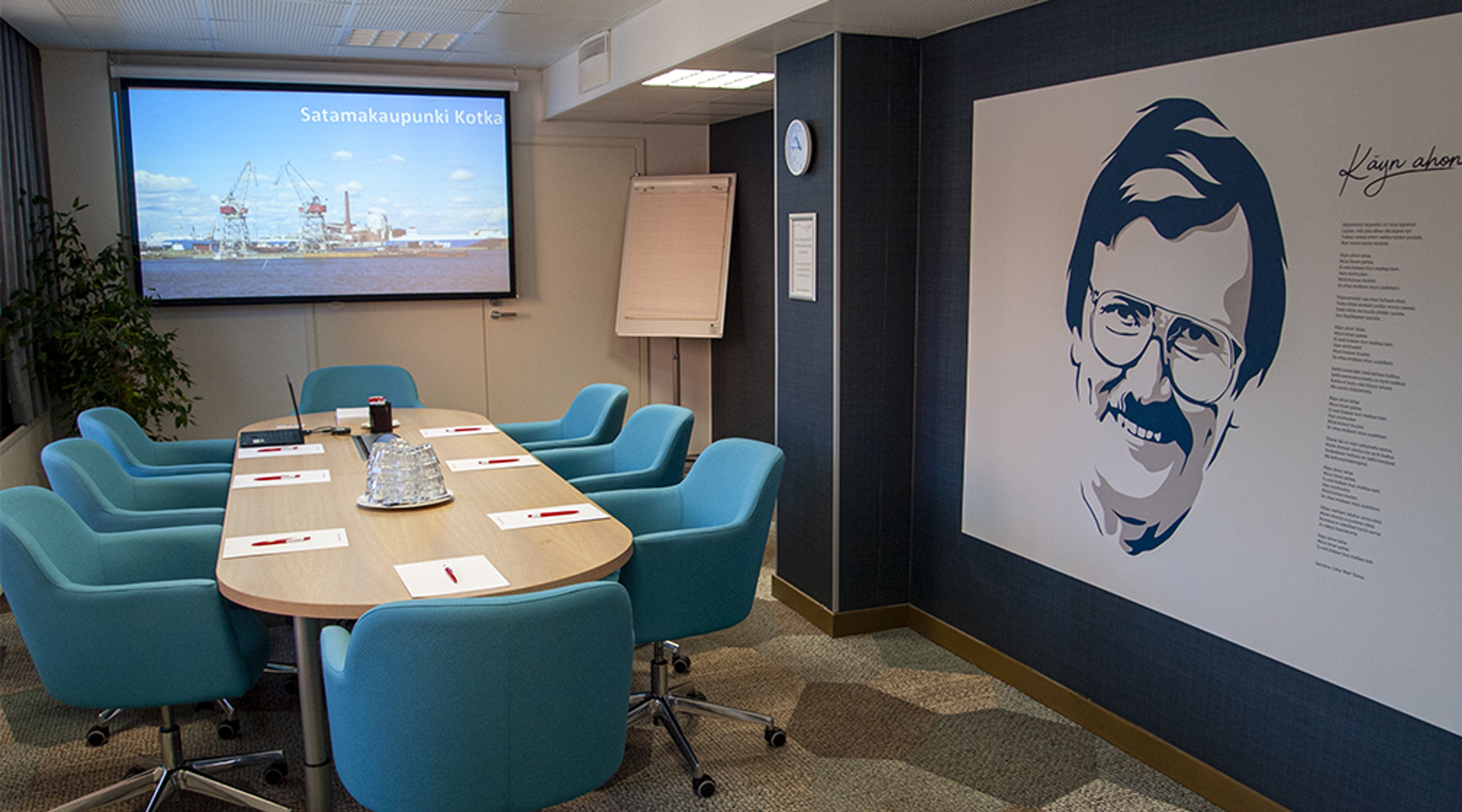 Conference room with wooden oval table and blue chairs. Large screen and flipchart on white wall, with decorative portrait on side wall.