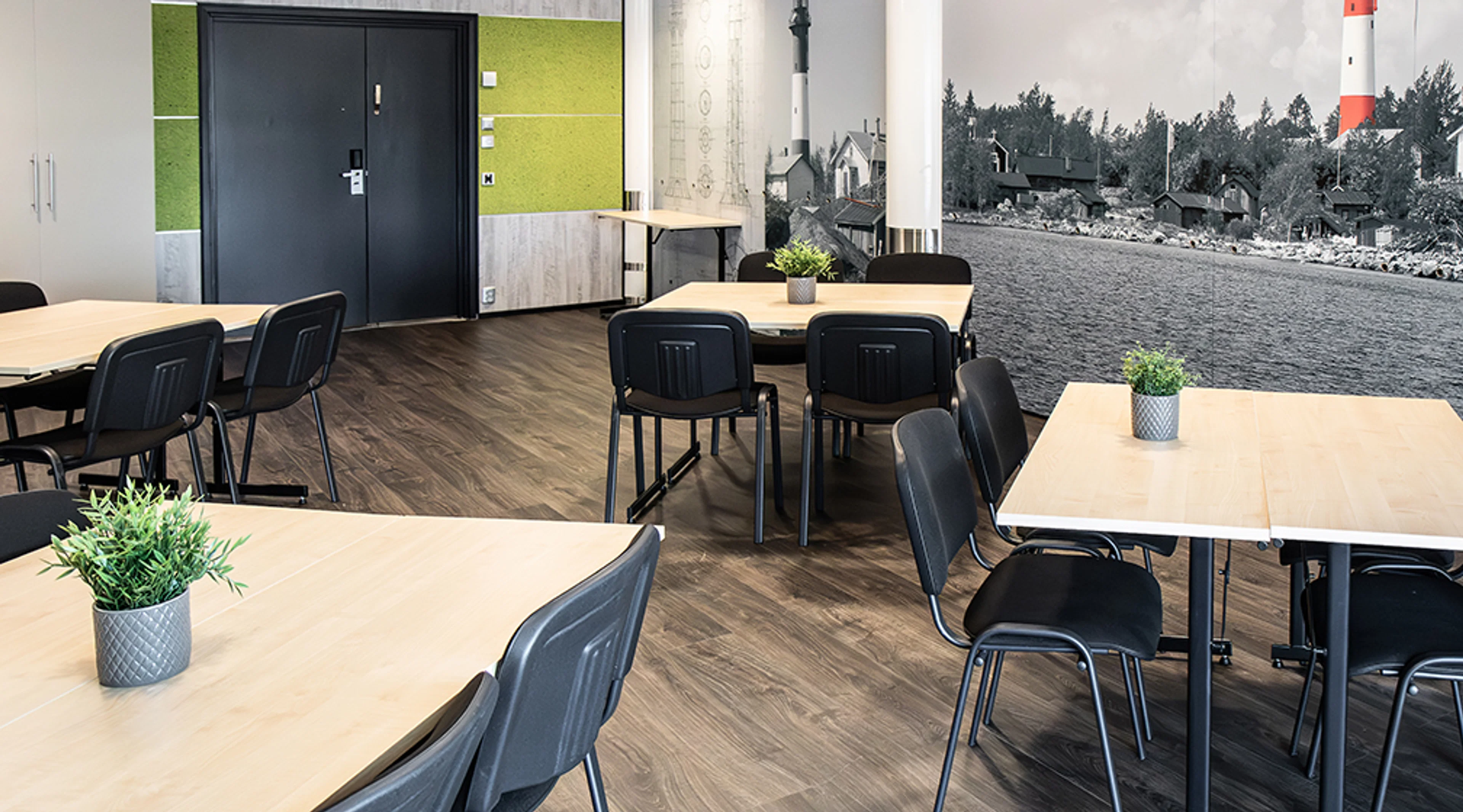 Meeting room with light wooden tables and black chairs. Wood-look laminate flooring, green acoustic wall, and black-and-white landscape wallpaper featuring a lighthouse.