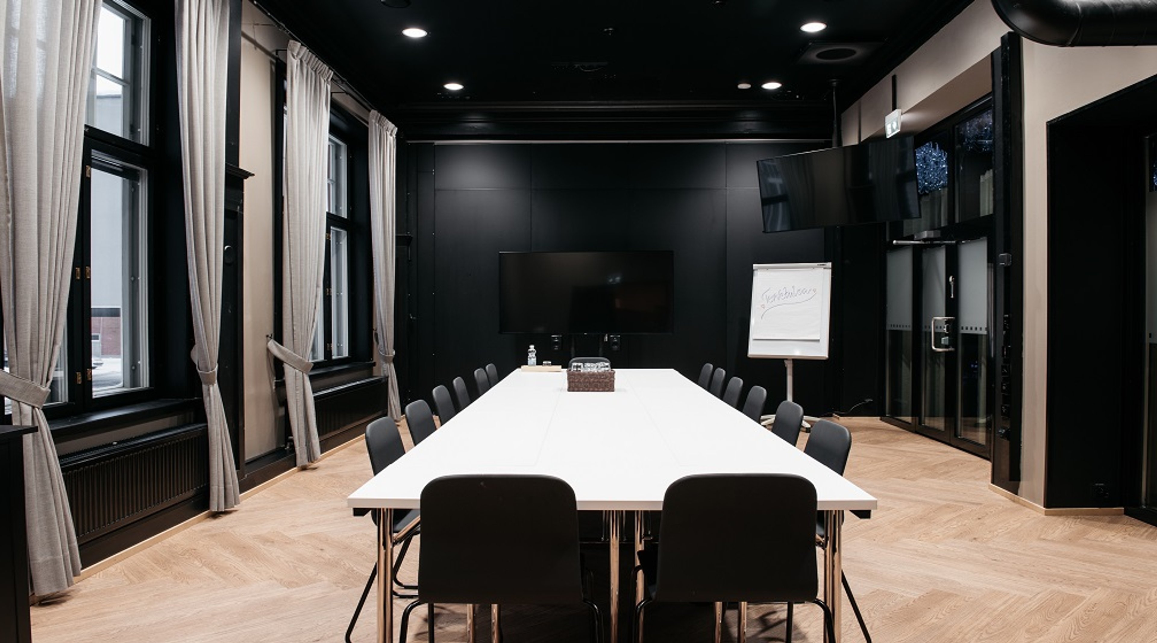 Modern conference room with black walls, large white table and black chairs. Tall windows with light curtains and wooden floor.