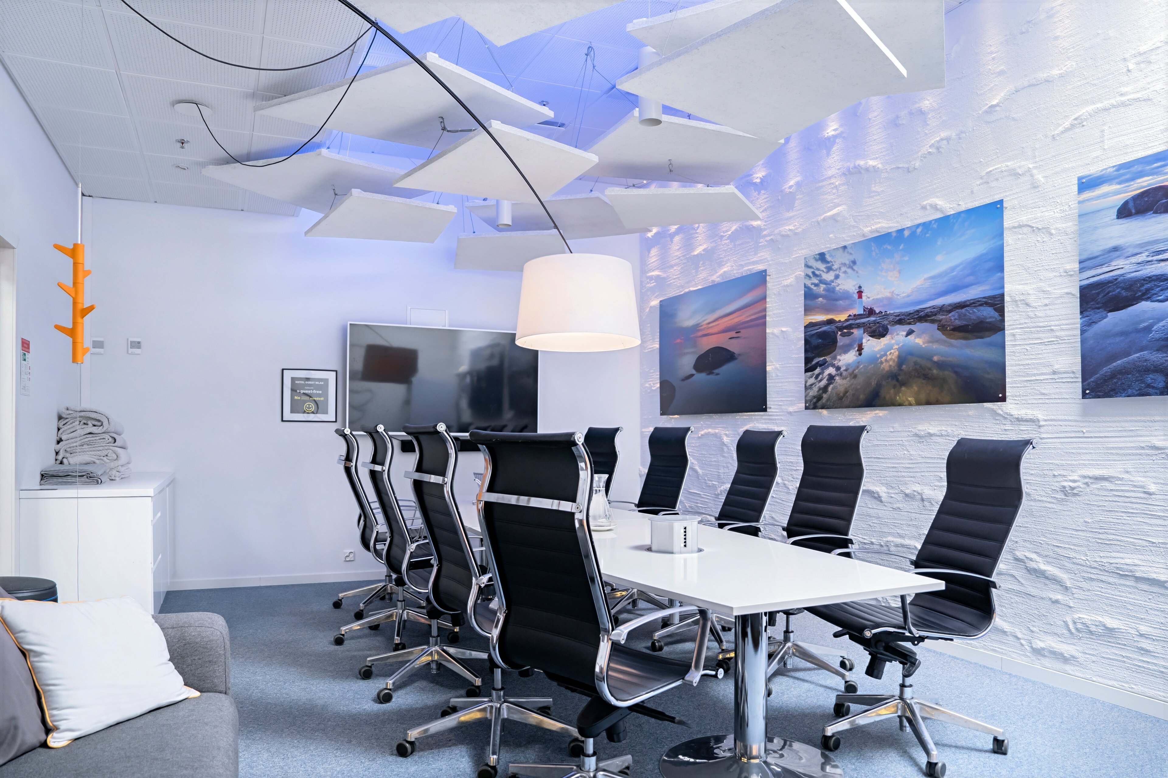 Modern white conference room with long table, black chairs and ocean landscape photos on the wall. White lamp and acoustic panels hang from the ceiling.