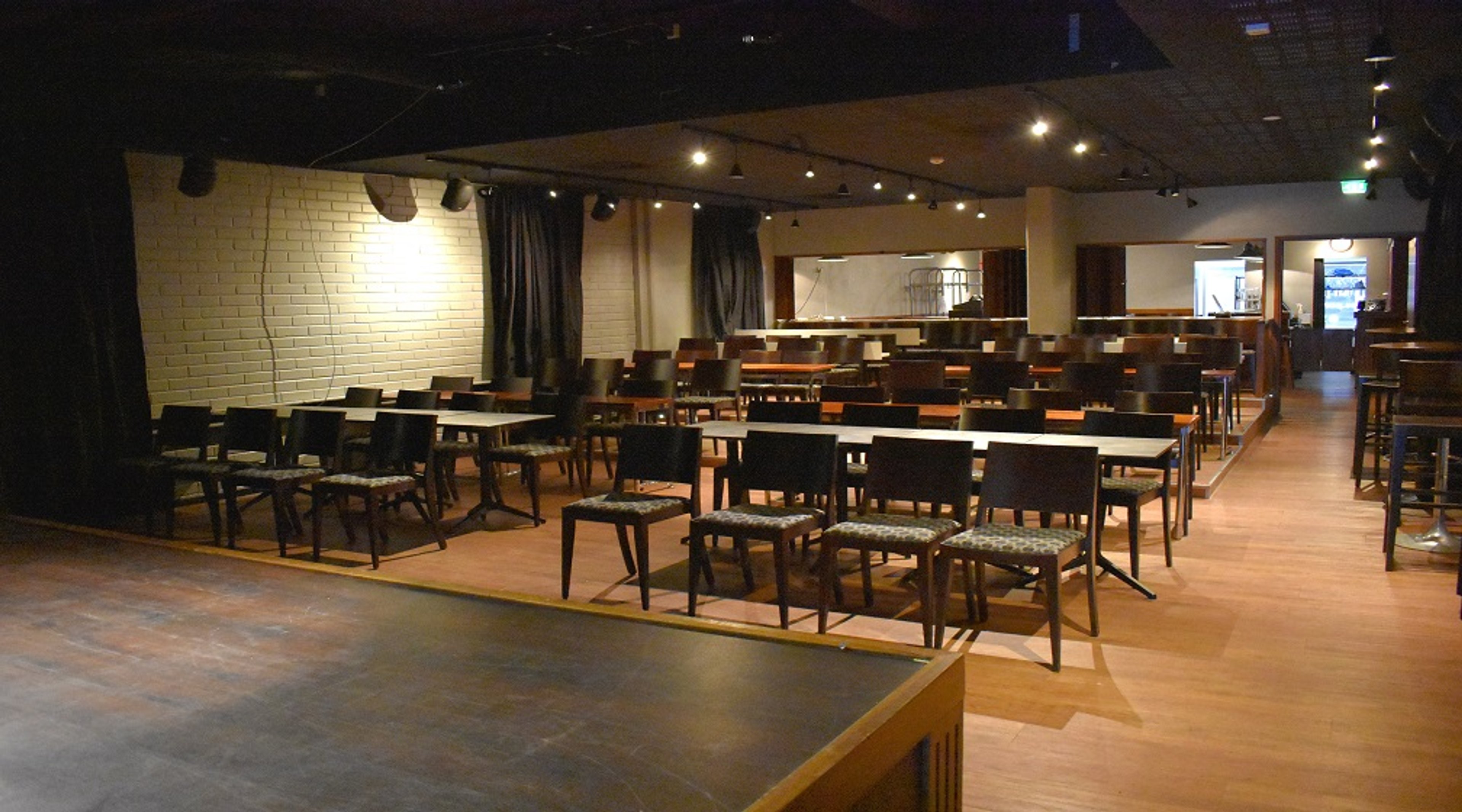Spacious venue with wooden floor, rows of chairs and tables, white brick wall and a small stage area in front. The space is well-lit.