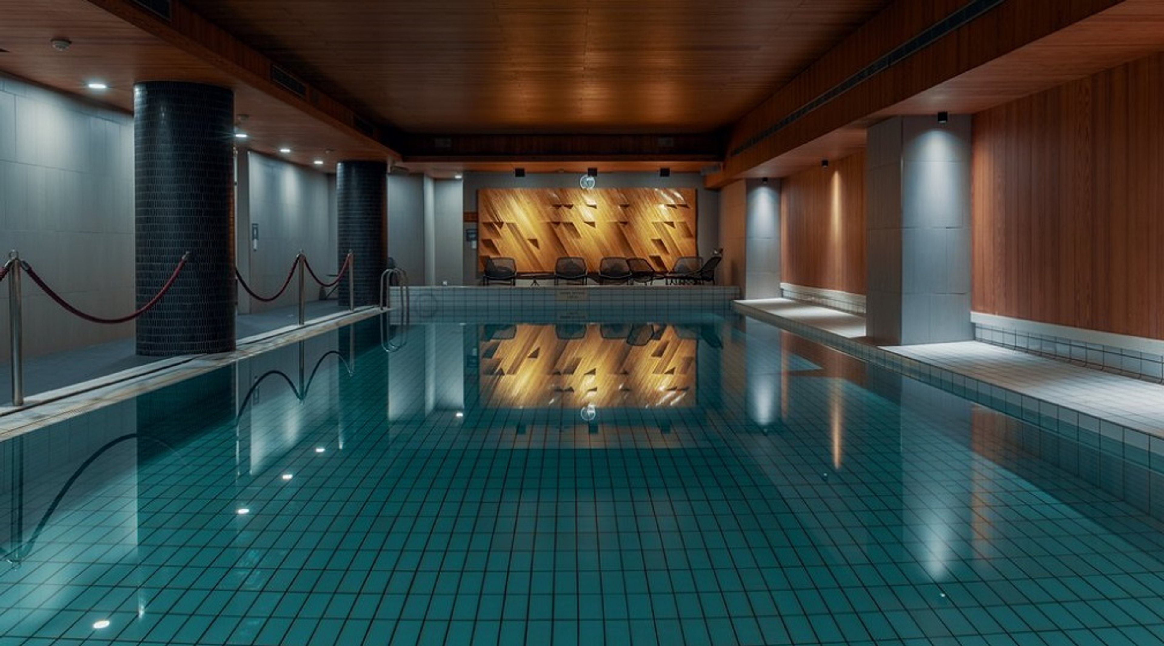 Fridfull atmosfär i en modern inomhuspool. Träpaneltak, stilfull belysning och turkosa plattor. Poolen har metallräcken.