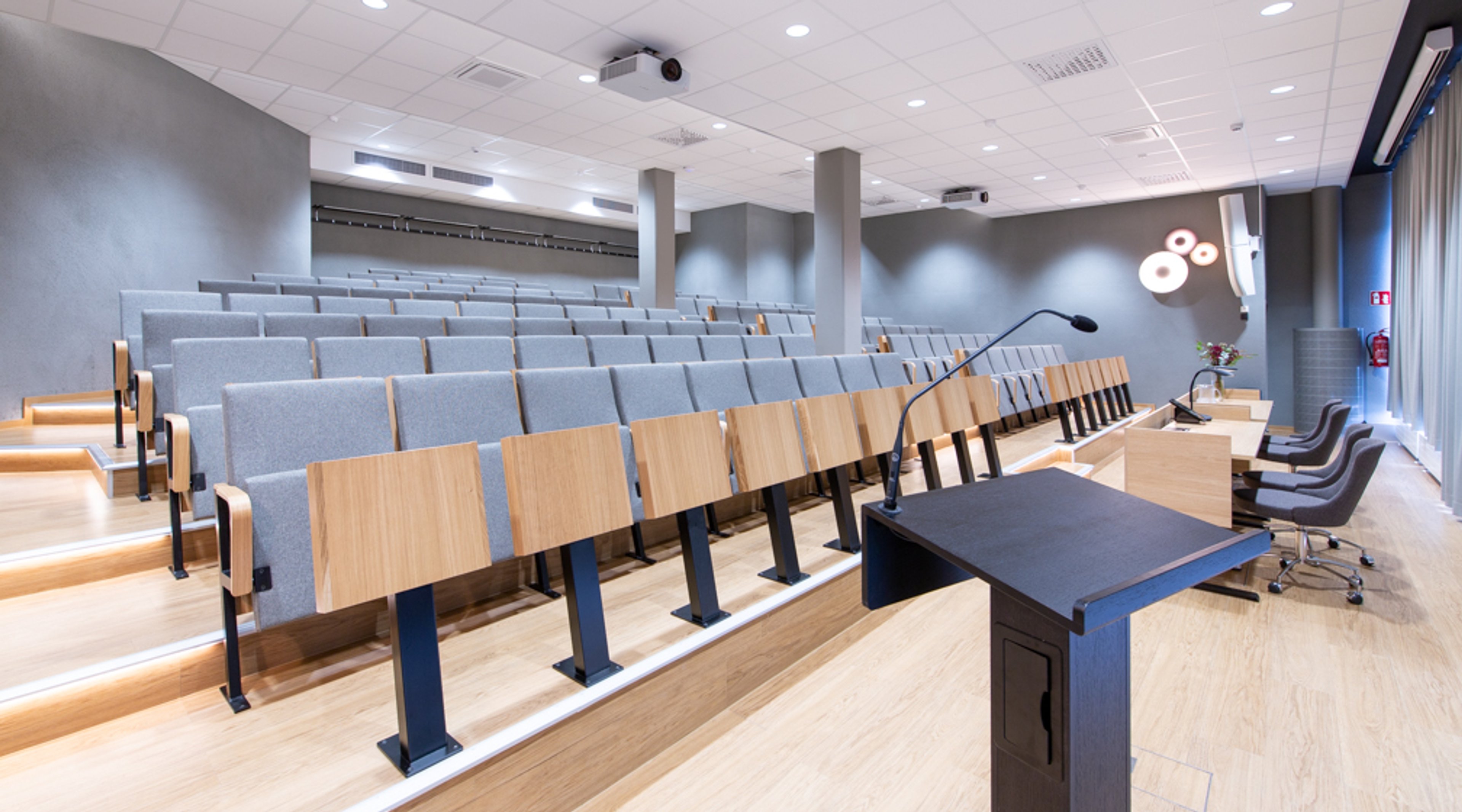 Modernt auditorium med blankpolerat trägolv, grå stoppade säten och träbord. Framtill finns en talarstol och kontrollbord.