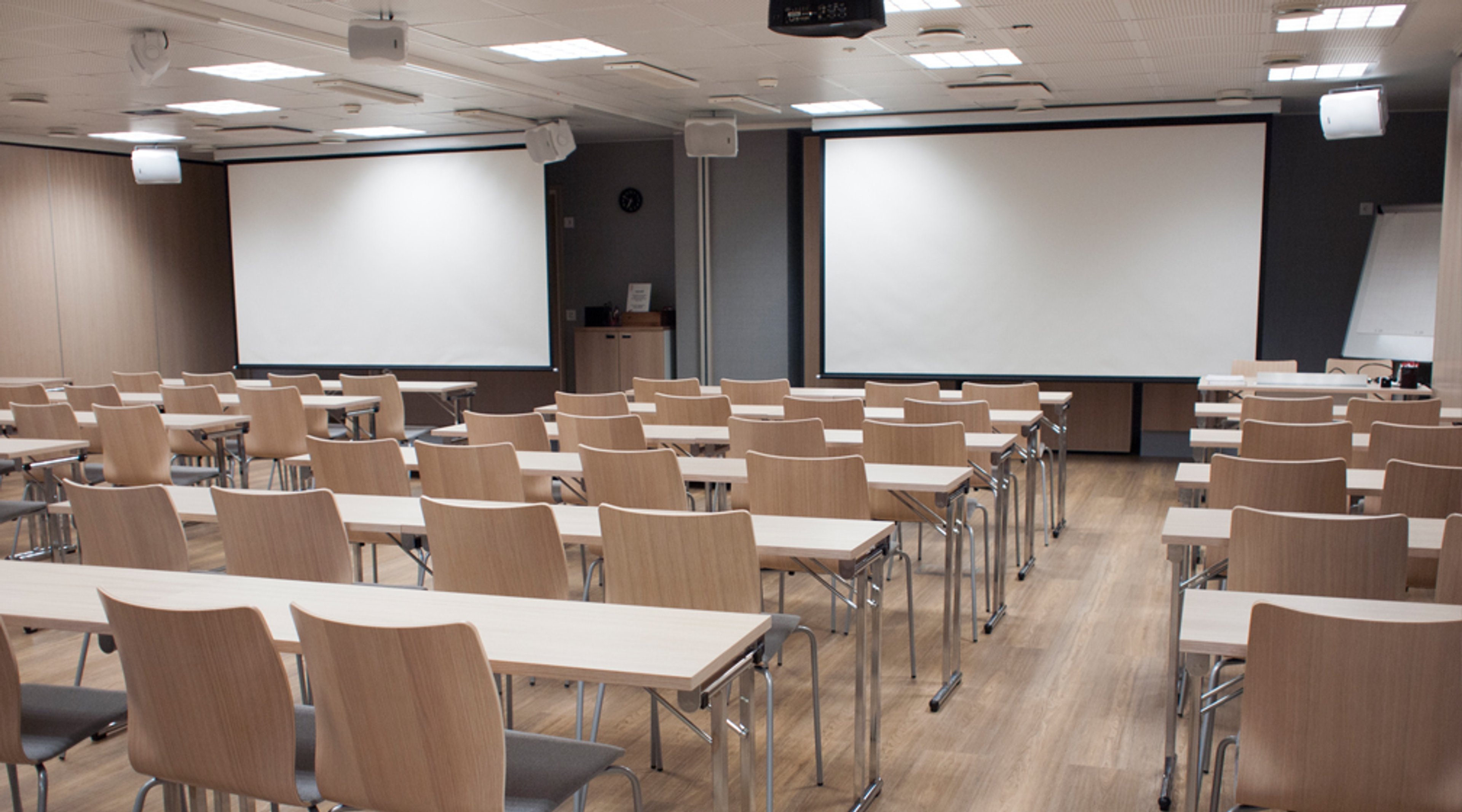Modern auditorium with light wooden flooring, wooden chairs and tables arranged in rows. Two white projection screens on the wall. Well-lit space.