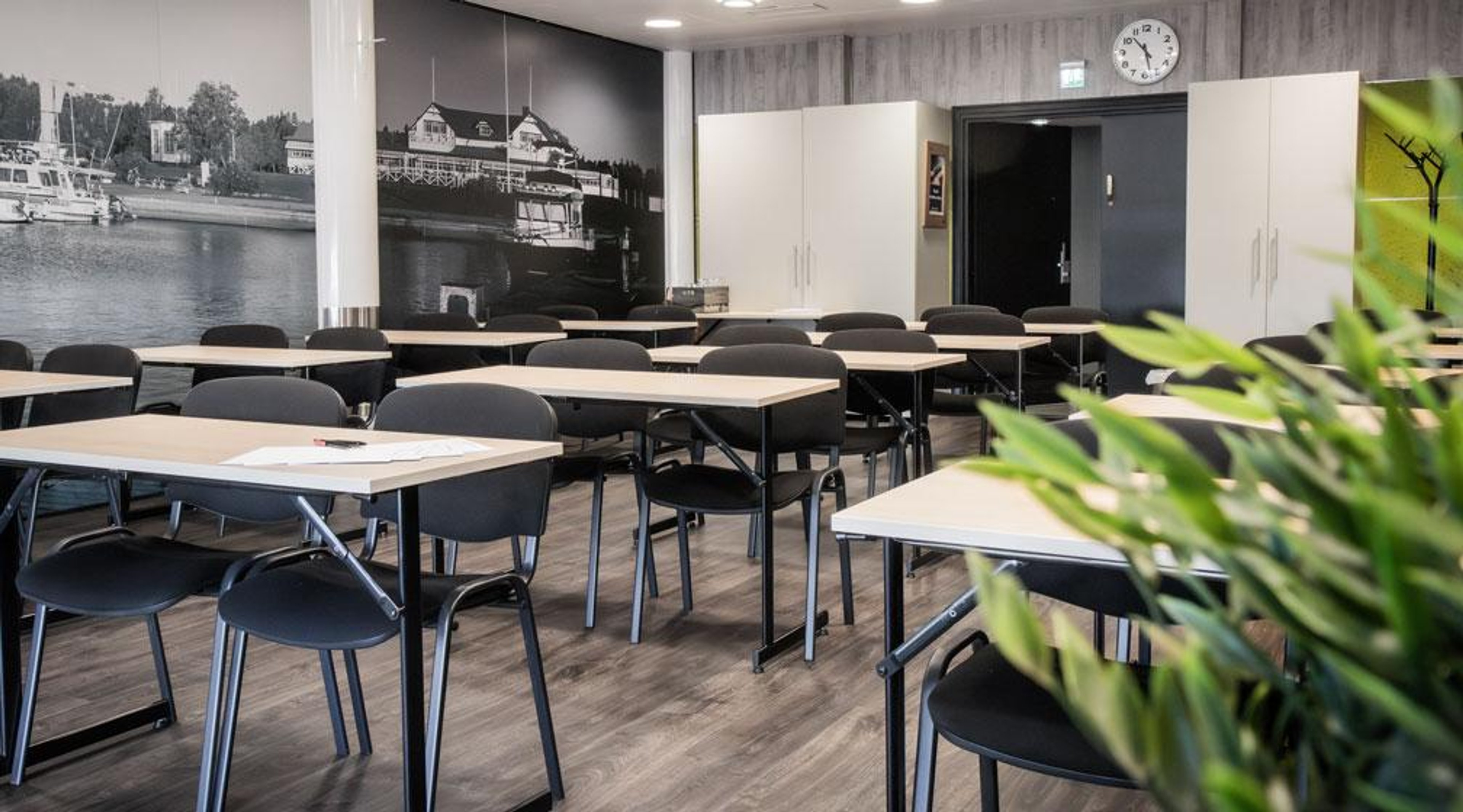 Training room with light-colored tables and black chairs. Background features a harbor photo mural and hard flooring.