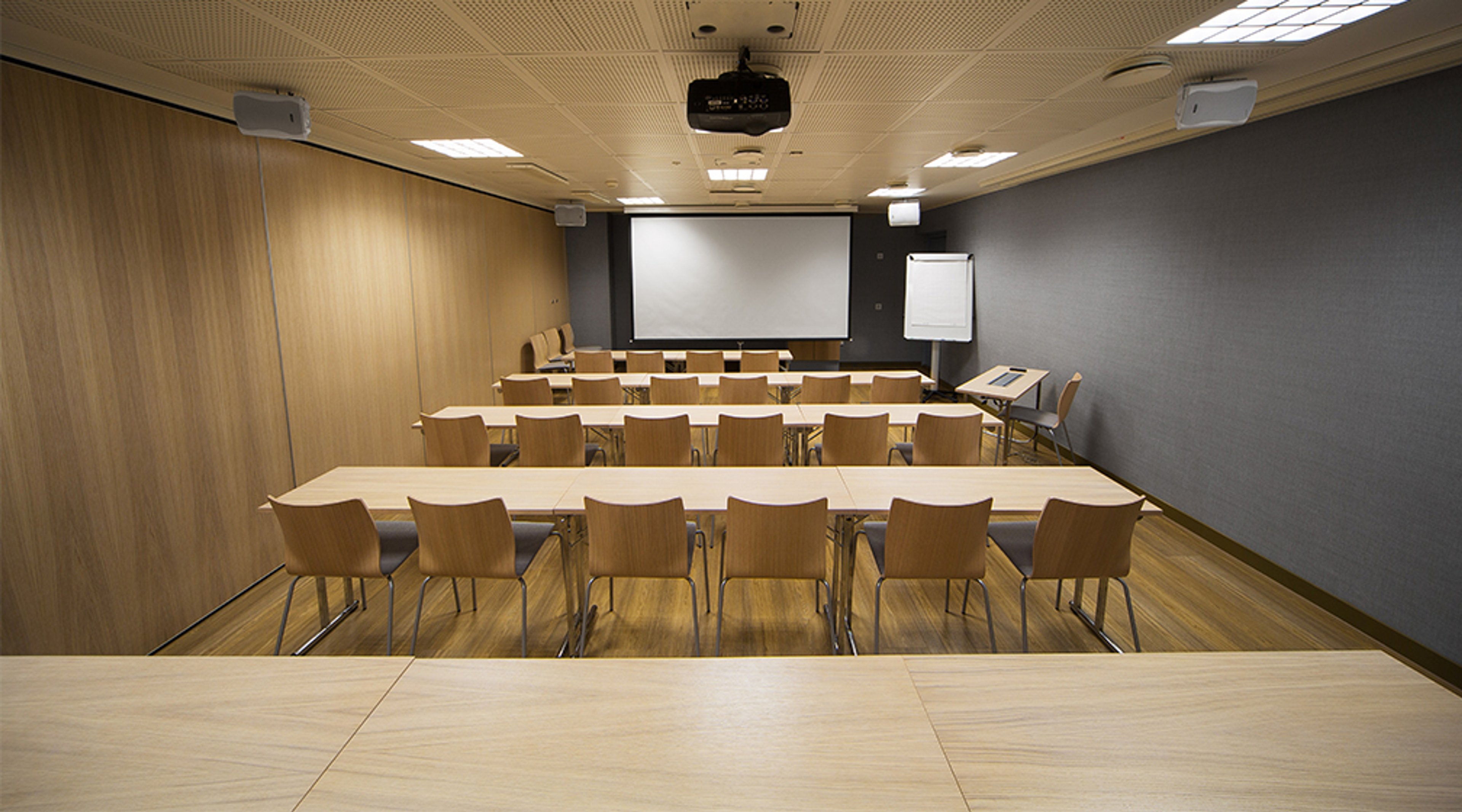 Conference room with wooden tables and chairs arranged in rows facing a projection screen. Room features wood panels and grey walls.