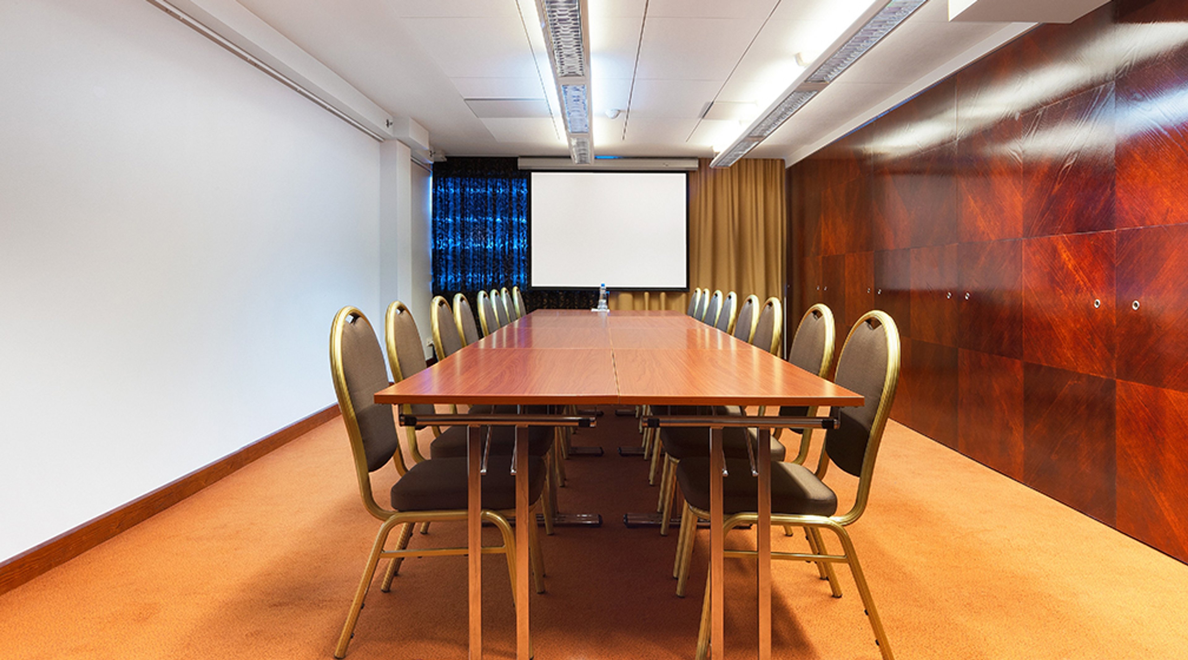 Conference room with long wooden table and chairs. Orange carpet, wooden panel wall, projection screen. Bright lighting and level floor.