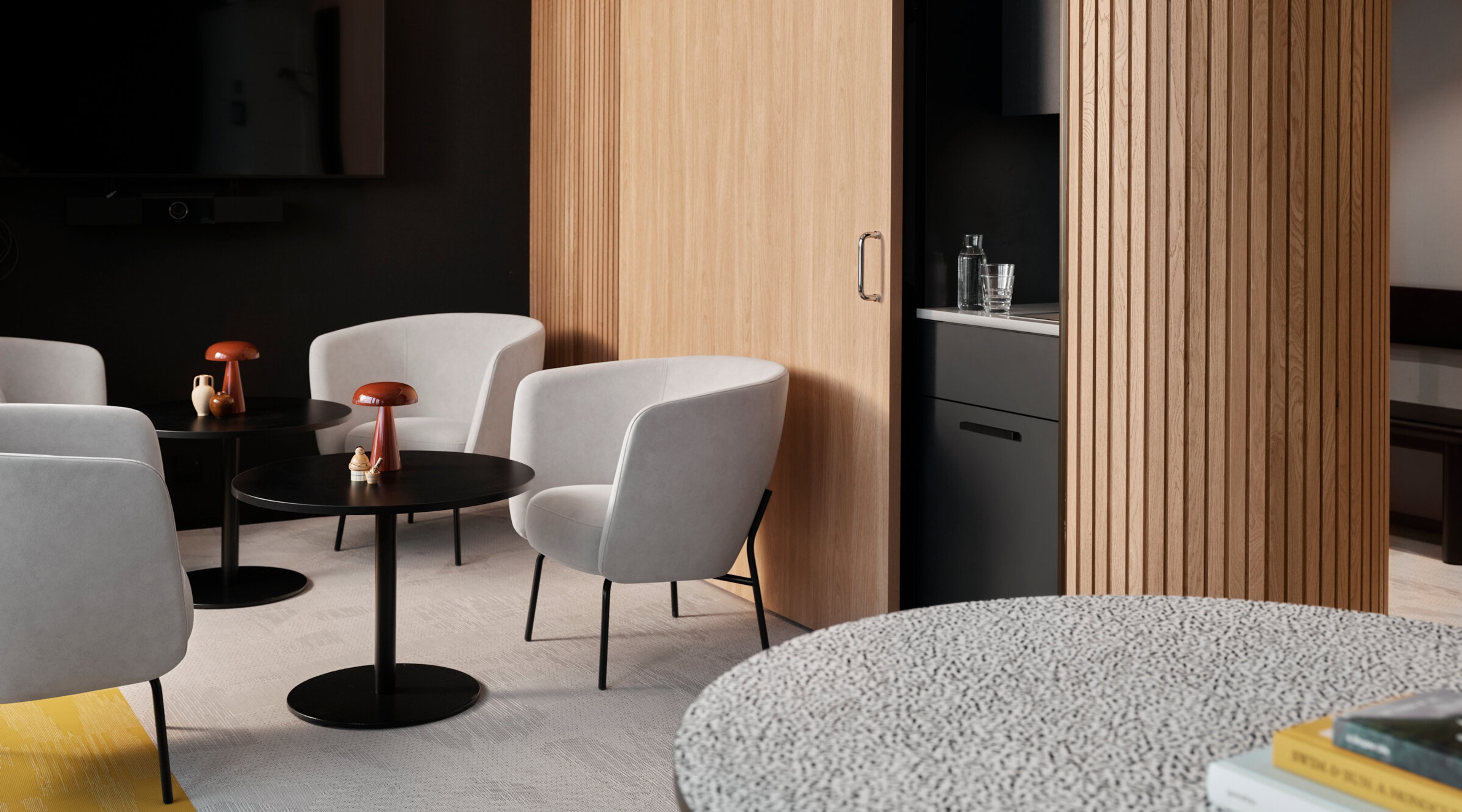 Modern lounge area with white armchairs, black tables and wooden slatted wall. Level light grey carpet flooring and built-in mini kitchenette.