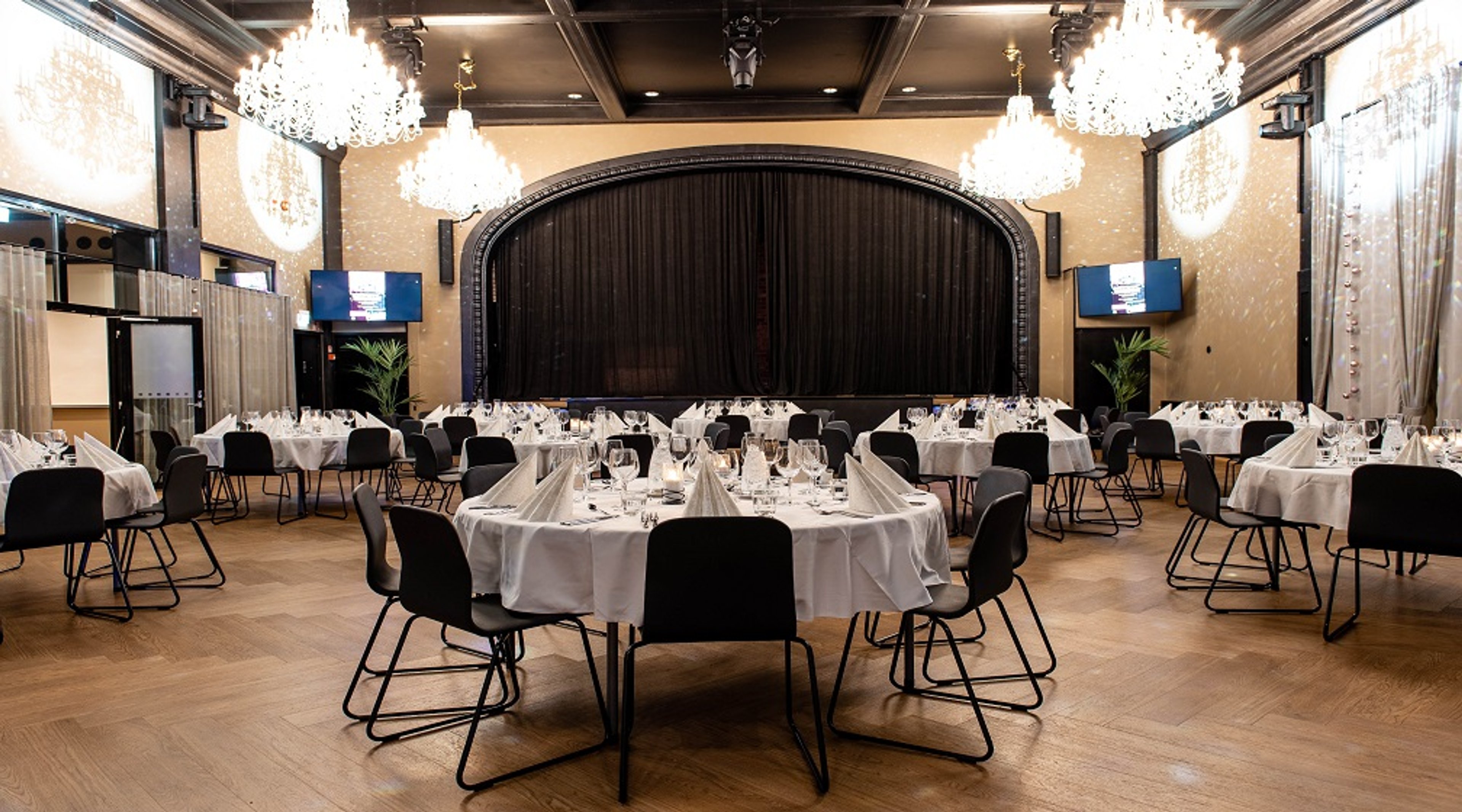 Banquet hall with round tables covered in white tablecloths and black chairs. Stage in background, crystal chandeliers overhead, wooden flooring.