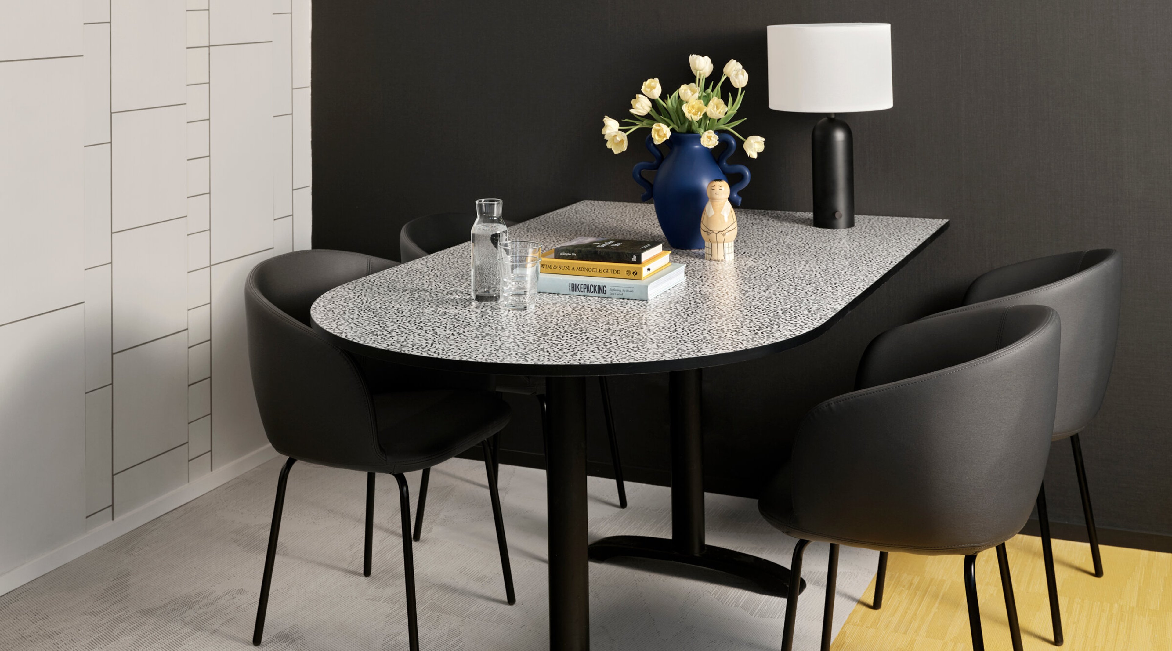 Modern dining area with terrazzo table, four black chairs, blue vase with light tulips and black table lamp with light shade.