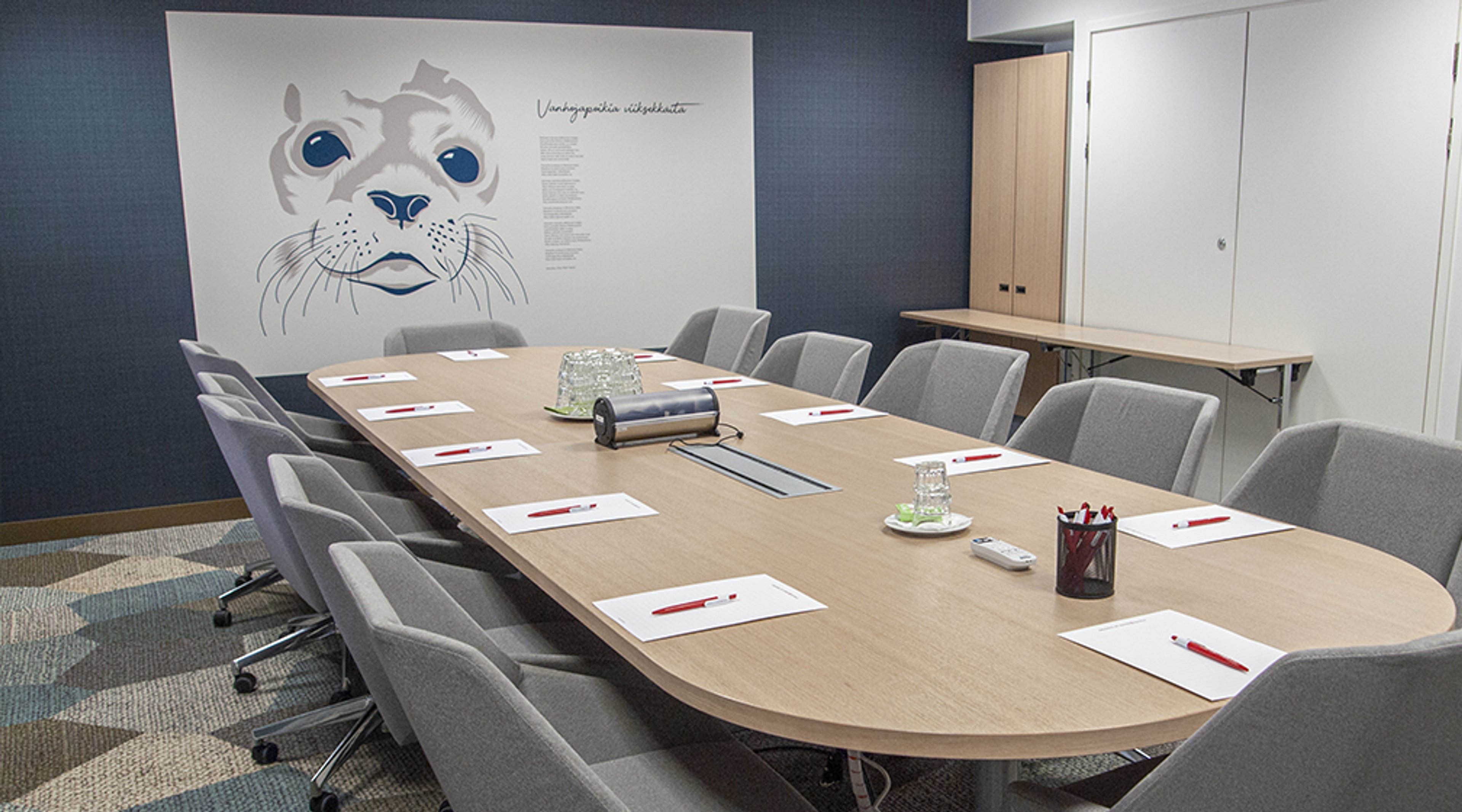 Meeting room with wooden table, grey chairs and seal face artwork on wall. Papers, pens and drinking glasses arranged on the table.